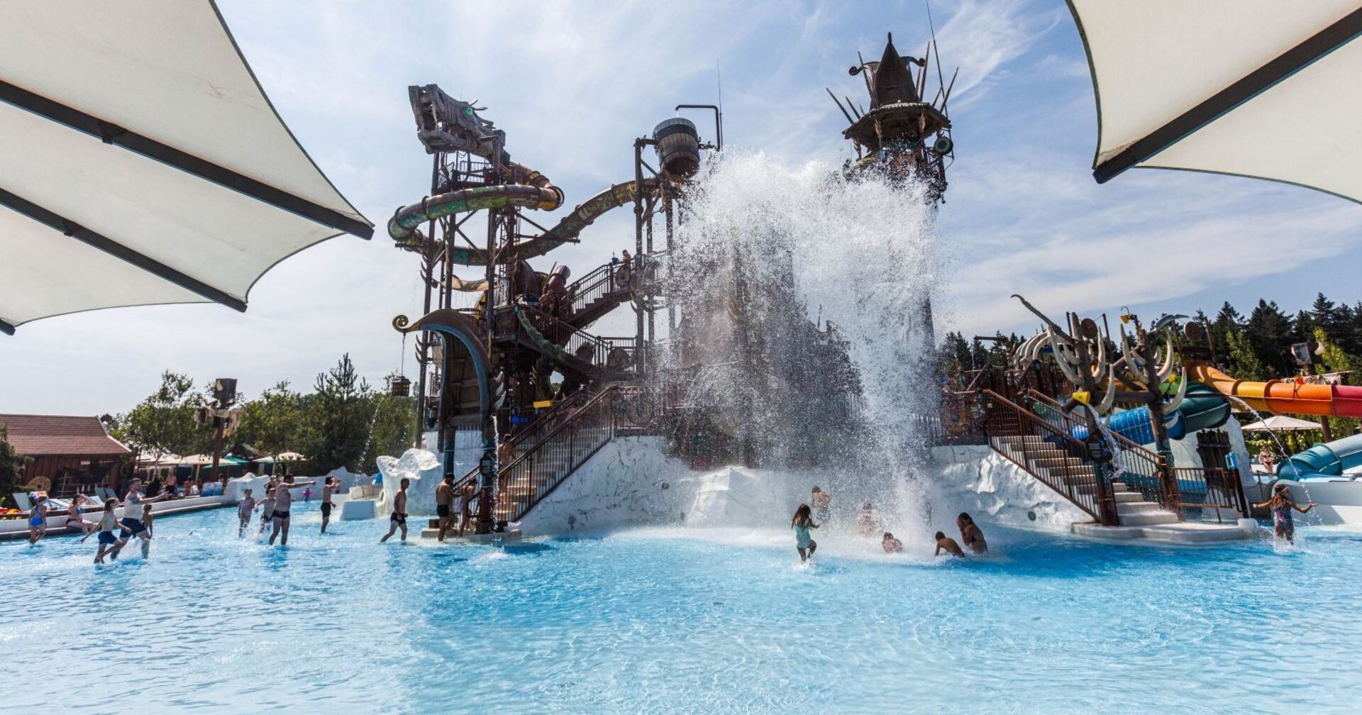 Das Bild zeigt einen großen Außenbereich eines Wasserparks mit mehreren Wasserrutschen und Spielanlagen im Hintergrund. Im Vordergrund befindet sich ein großes Becken, in dem gerade eine große Wassermenge aus einem Kipp-Eimer herabstürzt. Mehrere Kinder und Erwachsene spielen und baden im Wasser. Über dem Becken spenden weiße Sonnensegel Schatten. Im Hintergrund sind bunte Röhrenrutschen und weitere Wasserattraktionen zu sehen