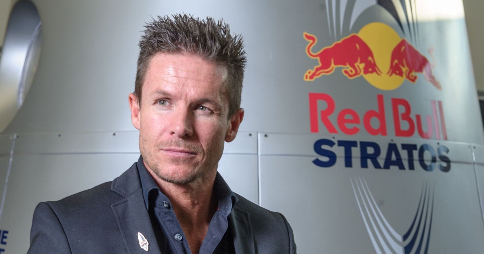 Felix Baumgartner steht in einem dunklen Anzug vor der Red Bull Stratos-Kapsel mit dem Red Bull Logo im Hintergrund.