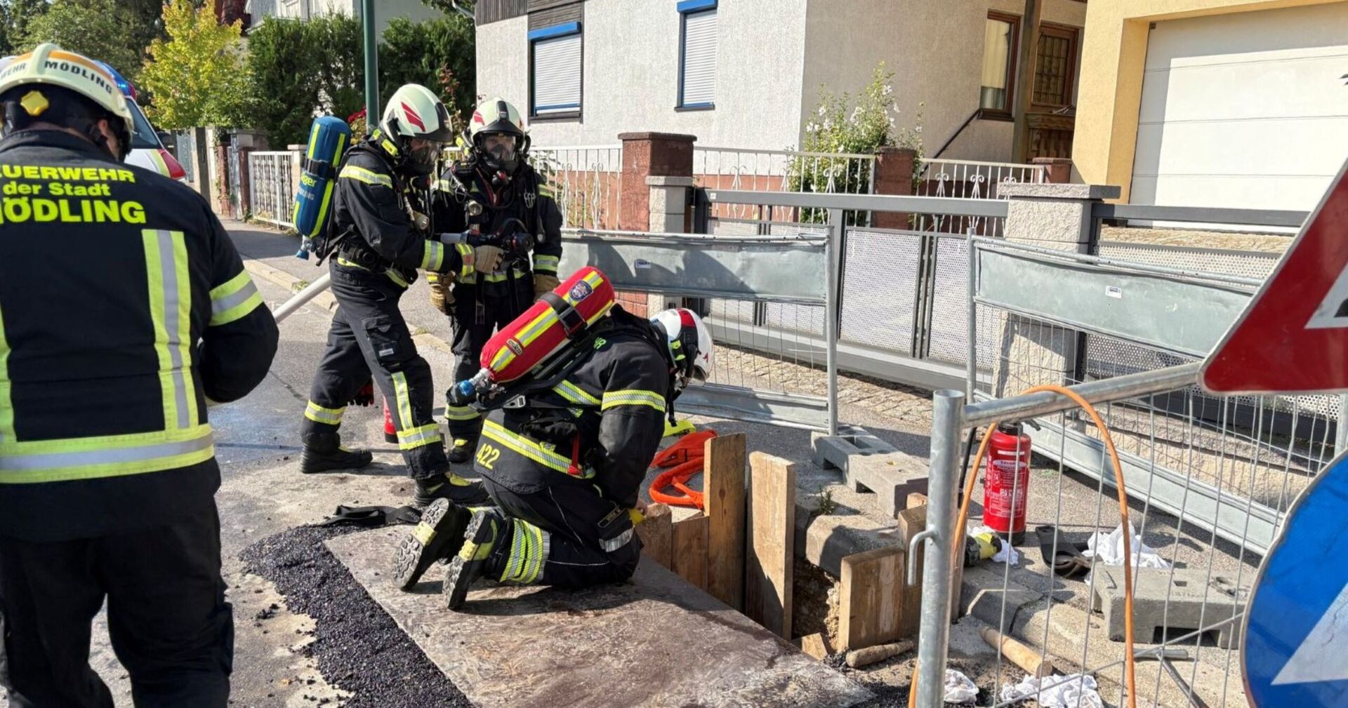 Mehrere Feuerwehrleute in Schutzkleidung und mit Atemschutz arbeiten an einer abgesicherten Baugrube auf einer Straße in Mödling nach einem Gasaustritt.