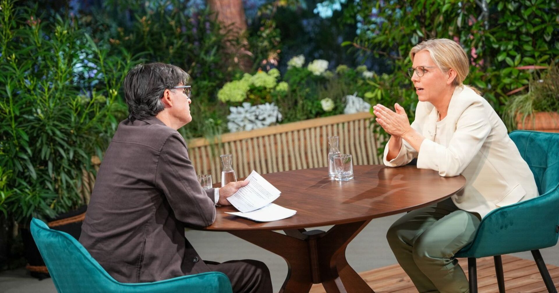 Klaus Webhofer und Leonore Gewessler sitzen sich beim ORF-Sommergespräch an einem runden Holztisch gegenüber, beide im Gespräch vertieft, im Hintergrund grüne Pflanzen.
