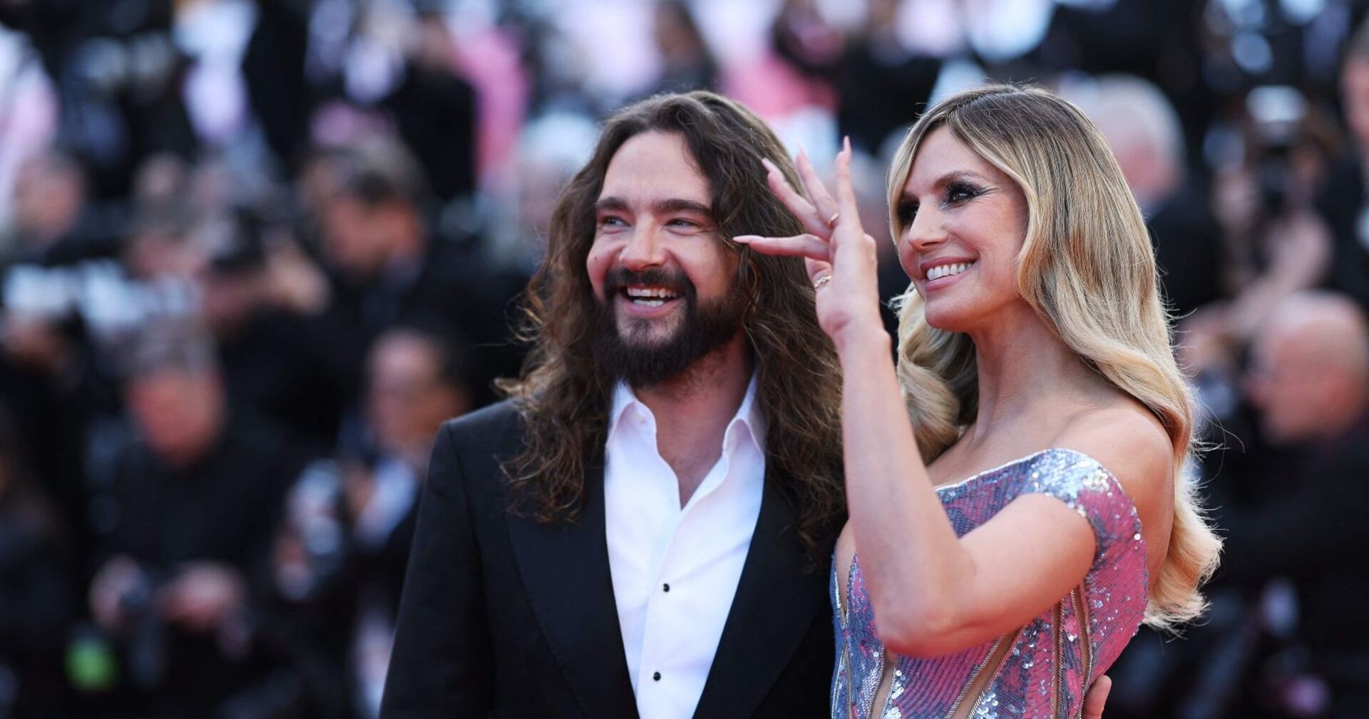 Heidi Klum und Tom Kaulitz posieren Arm in Arm auf dem roten Teppich. Klum trägt ein glitzerndes, schulterfreies Kleid und winkt lächelnd in die Menge, während Kaulitz in einem schwarzen Anzug lacht. Im Hintergrund sind zahlreiche Fotografen zu sehen.
