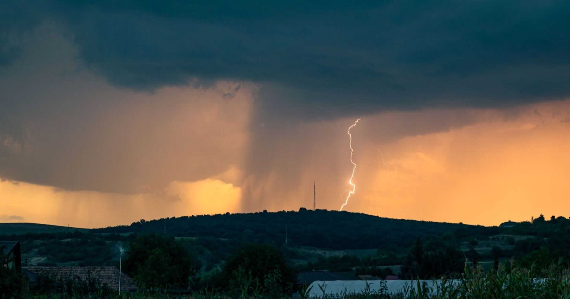 Dunkle Gewitterwolken über einer Hügellandschaft, ein Blitz schlägt neben einem Funkturm ein, im Vordergrund sind Häuser und Felder zu sehen.