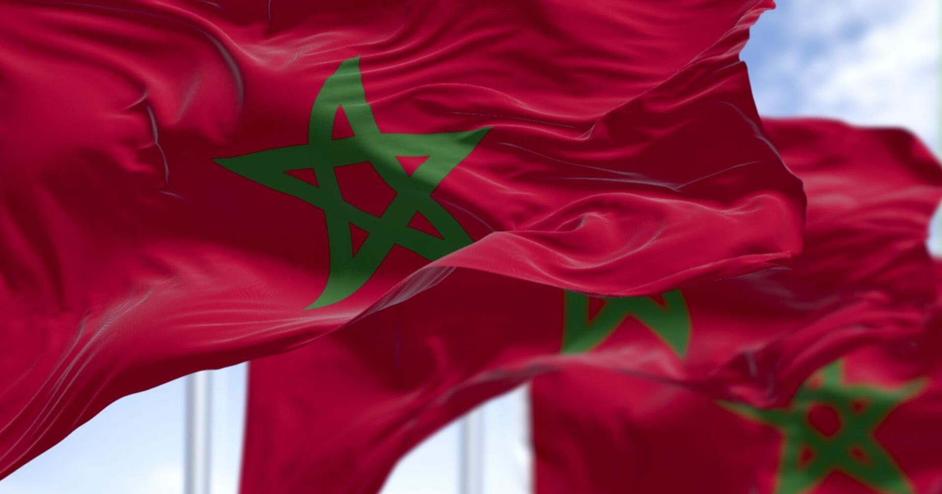 Nahaufnahme mehrerer wehender marokkanischer Flaggen mit grünem fünfzackigen Stern auf rotem Hintergrund vor blauem Himmel.