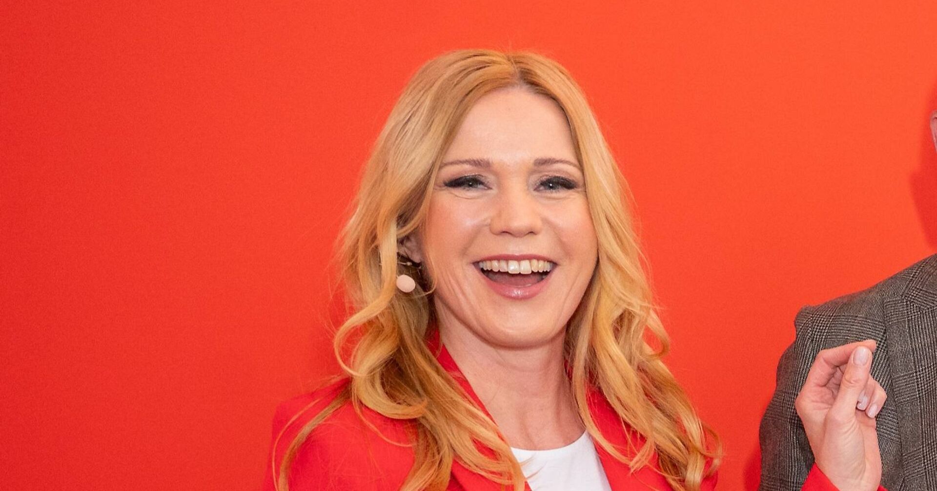Moderatorin Johanna Setzer, mit langen blonden Haaren lächelt in rotem Blazer vor rotem Hintergrund.
