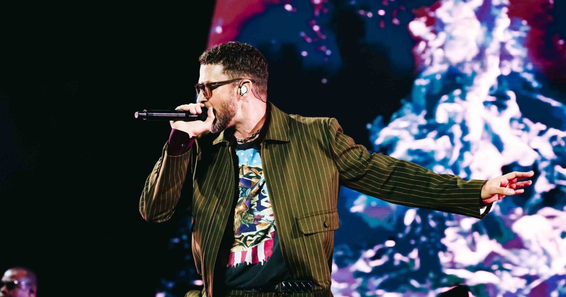 Justin Timberlake singt auf der Bühne, trägt ein gestreiftes Jackett und ein T-Shirt mit Grafikdruck, im Hintergrund eine farbige Lichtinstallation.
