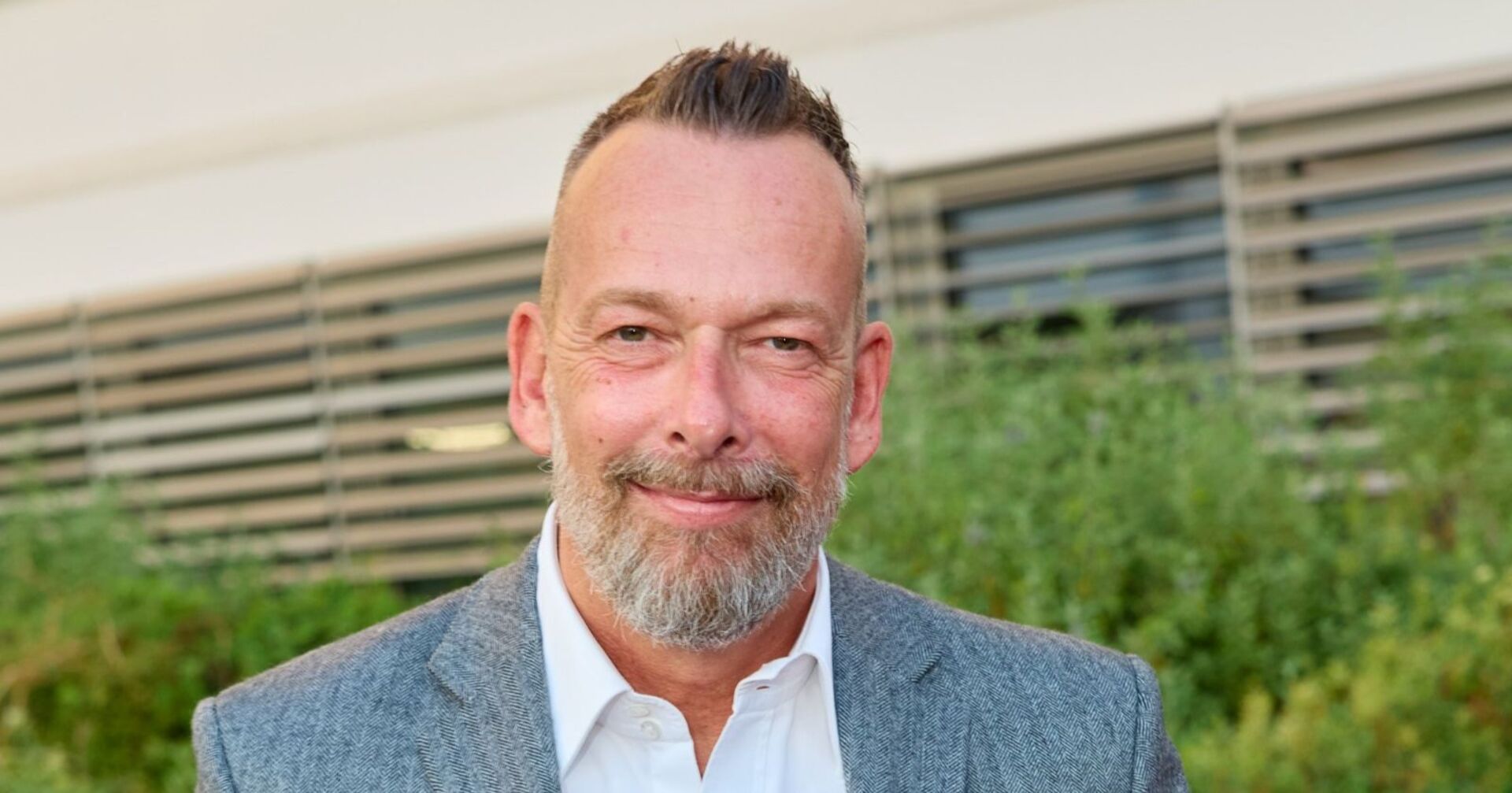 Porträt von Robert Kratky, der im grauen Sakko und weißem Hemd vor einem modernen Gebäude mit Lamellenfassade steht und in die Kamera lächelt.