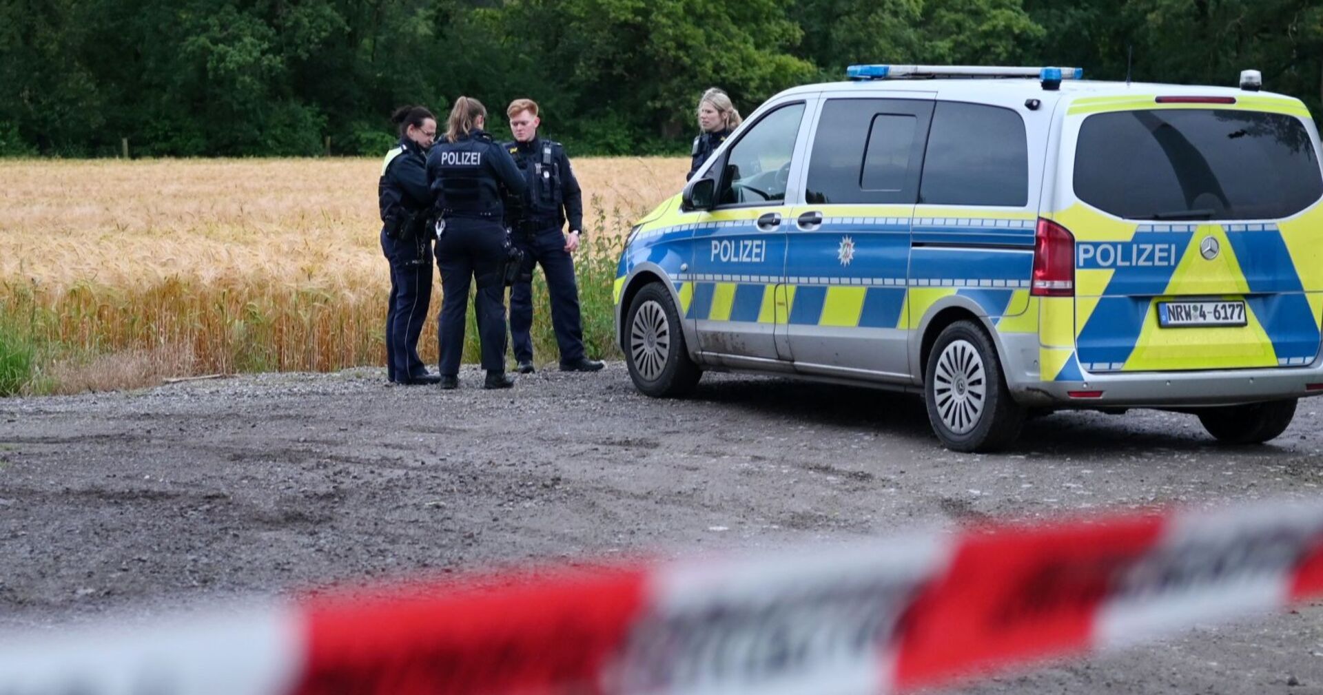 Mehrere Polizistinnen und Polizisten stehen neben einem Einsatzfahrzeug vor einem abgeernteten Feld, eine rot-weiße Absperrung ist im Vordergrund zu sehen.