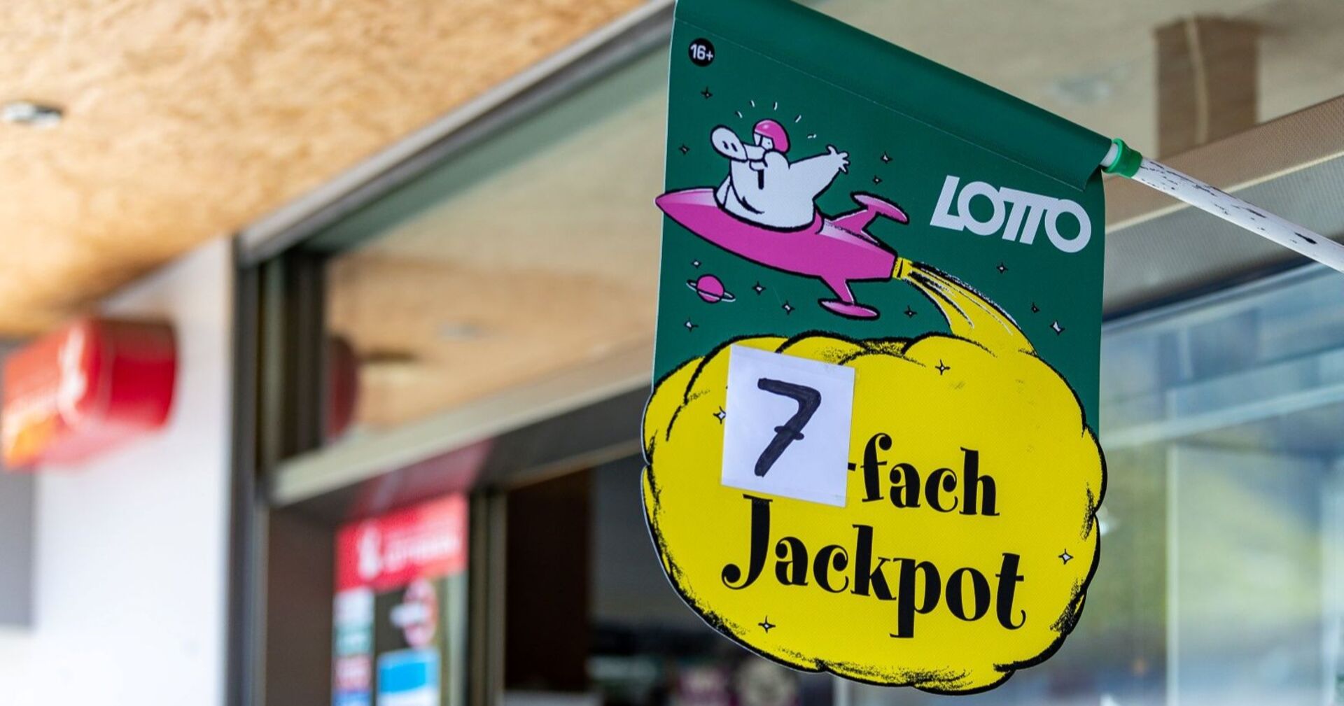 Grünes Lotto-Plakat mit gelber Wolke und der Aufschrift „7-fach Jackpot“ vor einer Trafik in Österreich.