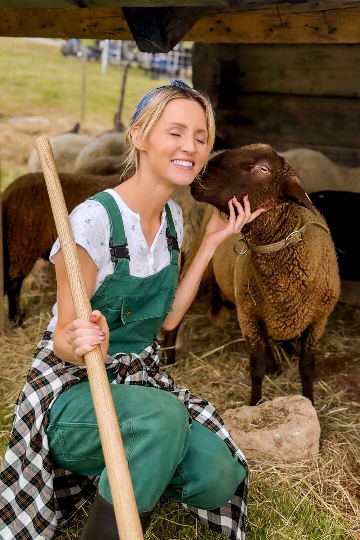 Melissa Naschenweng in grüner Latzhose und kariertem Hemd um die Hüfte, lächelnd beim Streicheln eines braunen Schafs während der Dreharbeiten auf einer Alm.