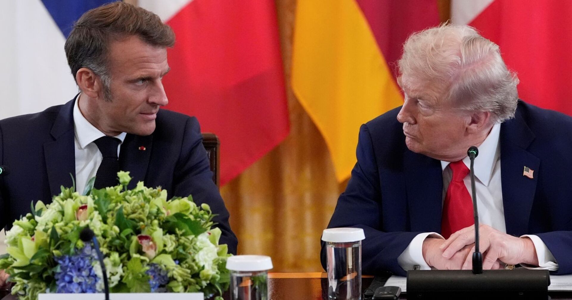 Frankreichs Präsident Emmanuel Macron und US-Präsident Donald Trump sitzen nebeneinander an einem Konferenztisch im Weißen Haus. Beide blicken sich ernst an, vor ihnen stehen Mikrofone, Gläser und ein großer Blumenschmuck. Im Hintergrund sind Flaggen verschiedener Länder zu sehen.
