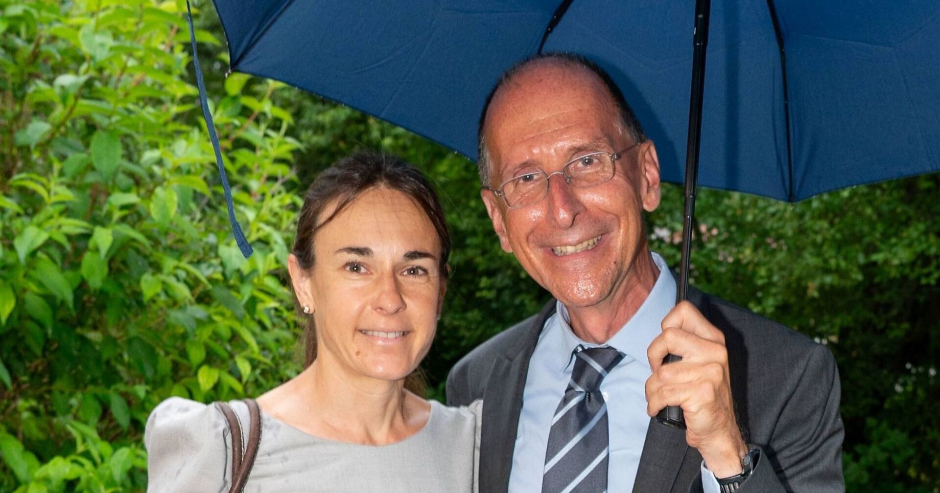 Peter Filzmaier und Carola Bendl lächeln gemeinsam unter einem blauen Regenschirm in die Kamera. Im Hintergrund ist grünes Blattwerk zu sehen.