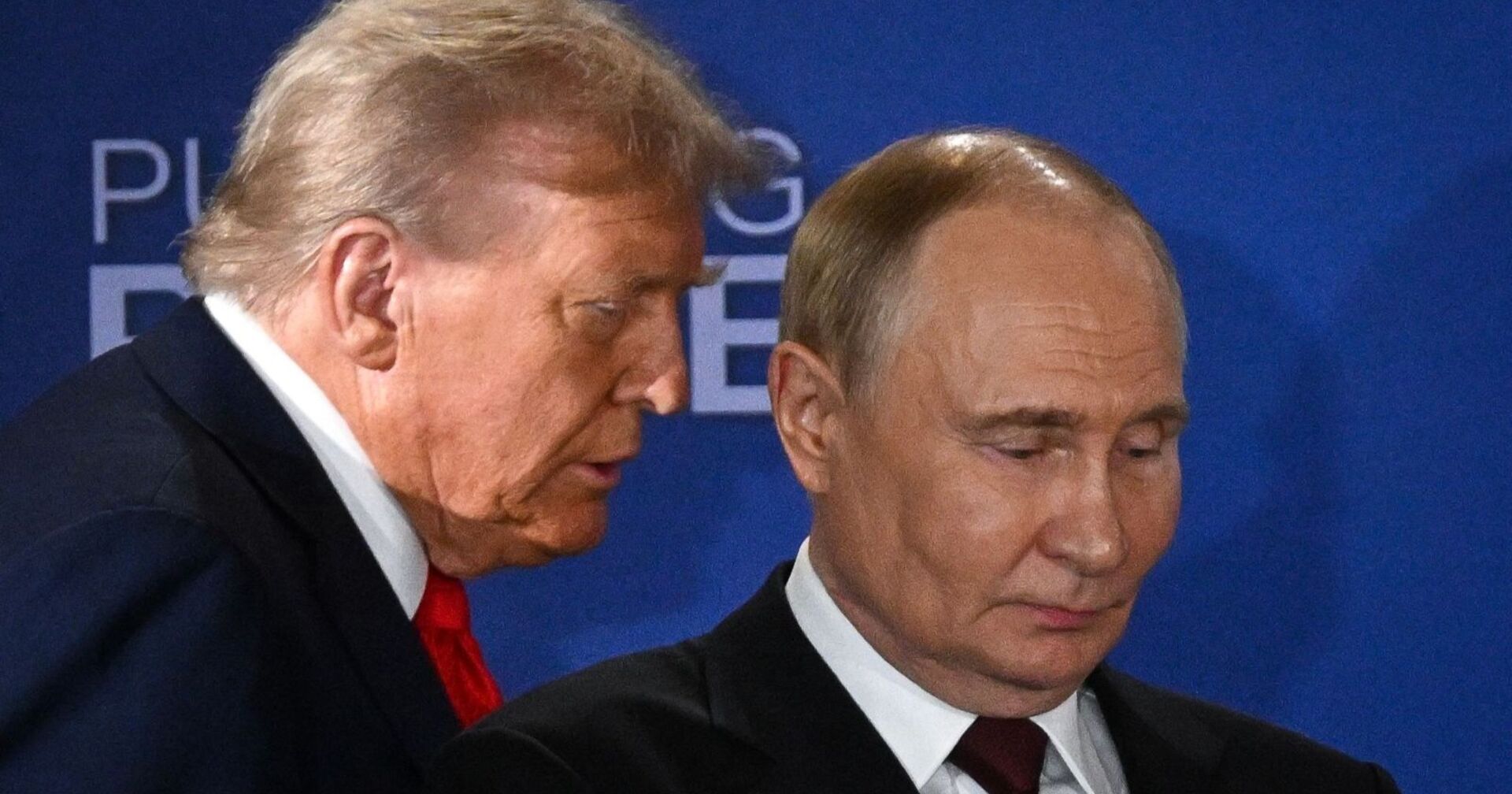 Donald Trump spricht dicht neben Wladimir Putin, der mit ernster Miene nach unten blickt. Beide tragen Anzug, im Hintergrund eine blaue Wand mit dem Schriftzug &quot;Pursuing Peace&quot;.