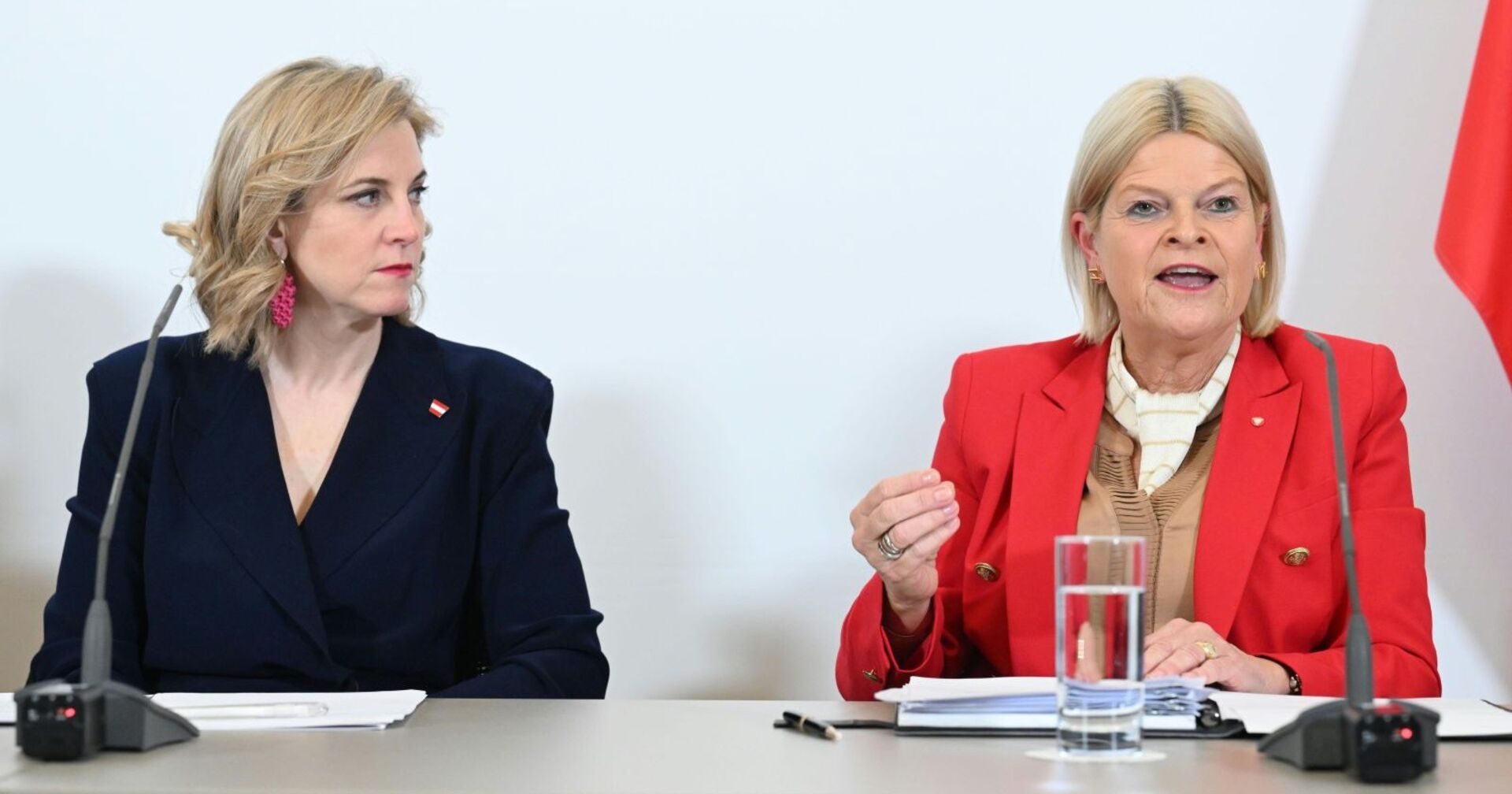Beate Meinl-Reisinger und Klaudia Tanner sitzen nebeneinander bei einer Pressekonferenz, sprechen vor Mikrofonen, im Hintergrund eine weiße Wand und rot-weiße Fahne.