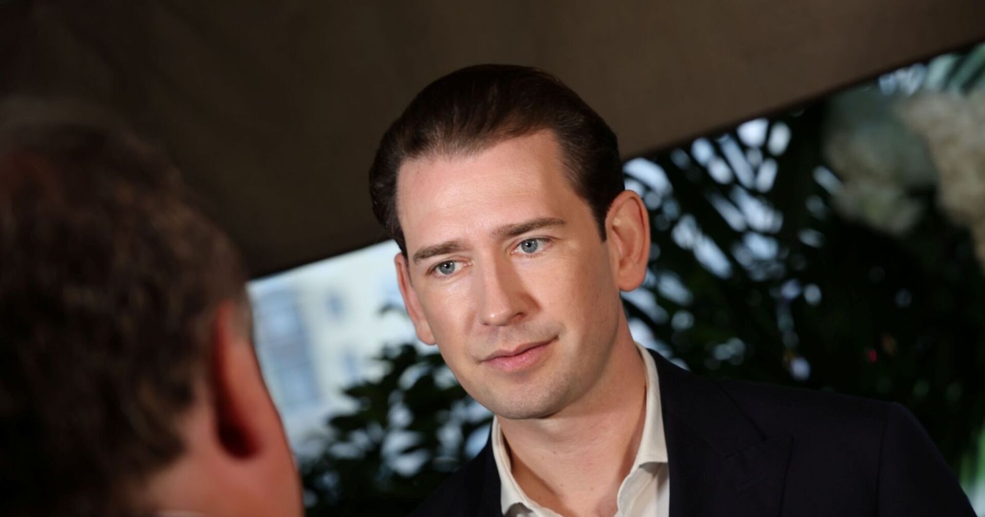 Sebastian Kurz im Anzug bei einem Interview, im Vordergrund eine weitere Person unscharf zu sehen, Hintergrund mit Pflanzen und unscharfer Architektur.