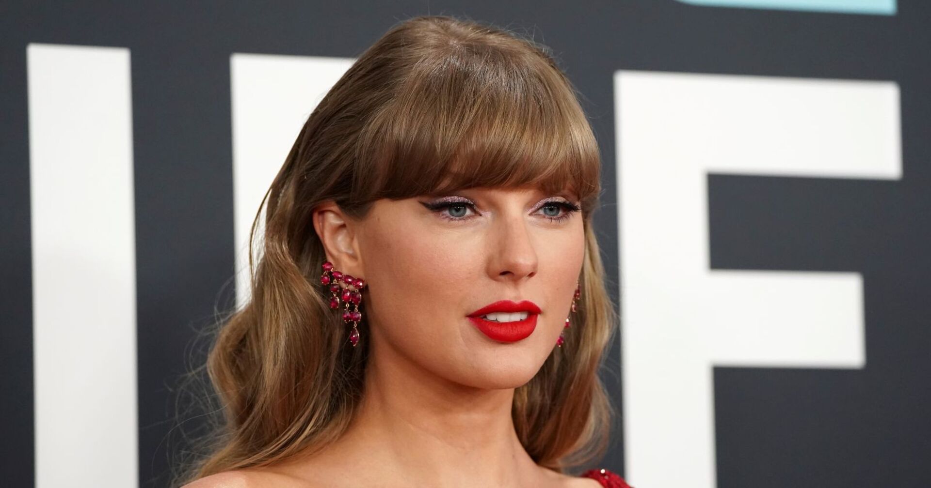 Nahaufnahme von Taylor Swift mit roten Lippen, funkelnden Ohrringen und glitzerndem Augen-Make-up vor schwarzem Hintergrund mit weißen Buchstaben.