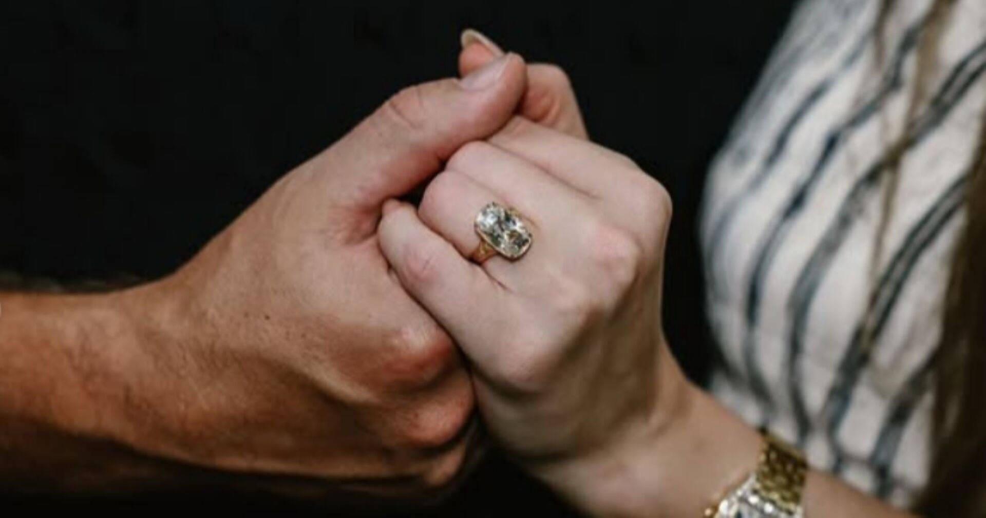 Nahaufnahme zweier sich haltender Hände, im Fokus ein großer rechteckiger Diamantring mit Gelbgoldfassung am Finger von Taylor Swift, die ein gestreiftes Kleid trägt.