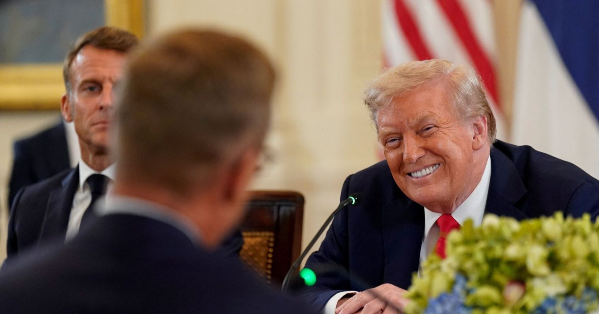 Donald Trump lächelt breit bei einem Treffen im Weißen Haus,  im Hintergrund Emmanuel Macron mit ernstem Blick.