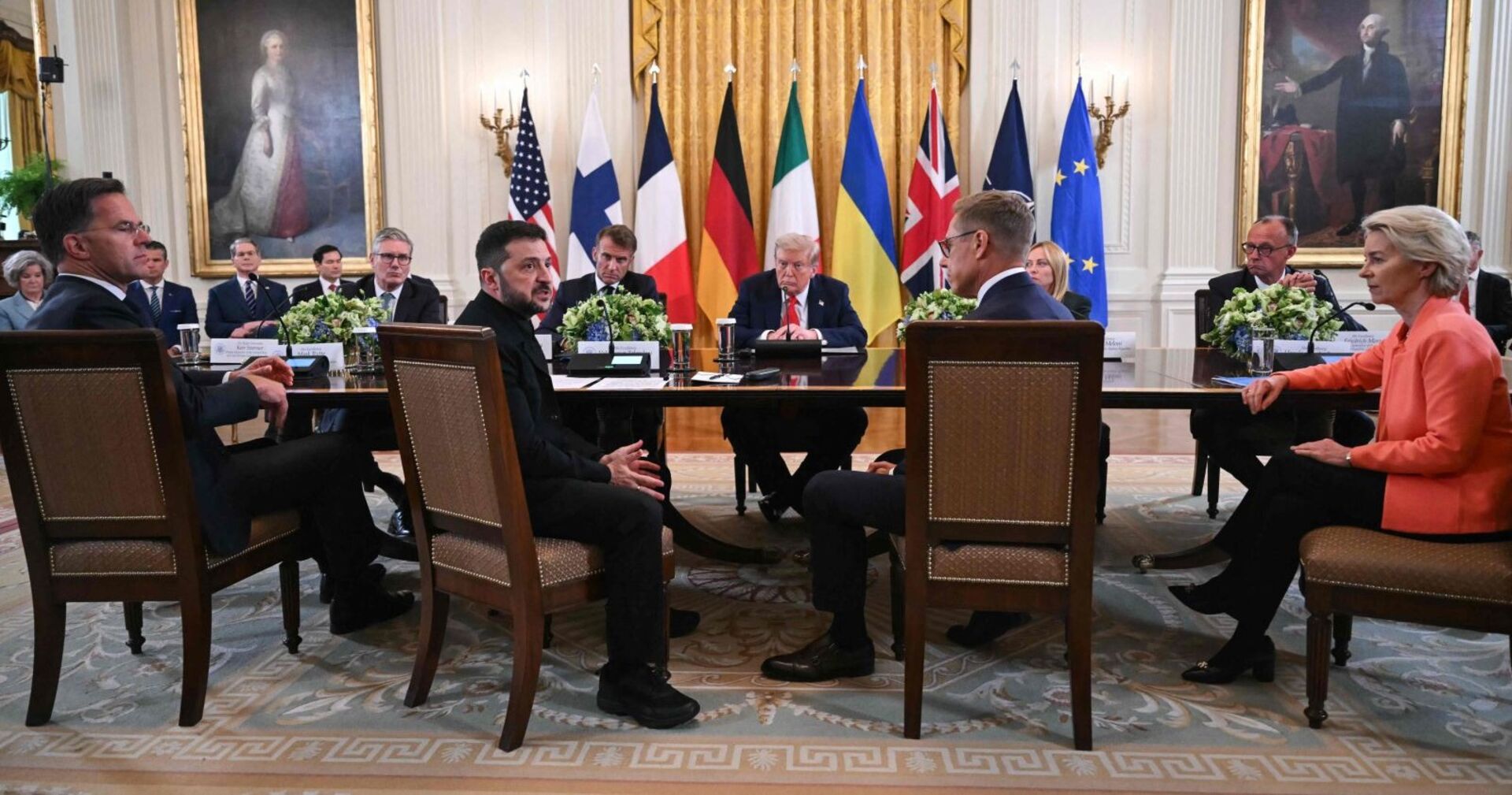 Ukraines Präsident Selenskyj, US-Präsident Trump und führende Politiker aus der EU und der NATO sitzen im Weißen Haus an einem großen Konferenztisch. Im Hintergrund stehen Landesflaggen, links und rechts hängen Porträts früherer Persönlichkeiten. Die Anwesenden blicken ernst, die Atmosphäre wirkt konzentriert.