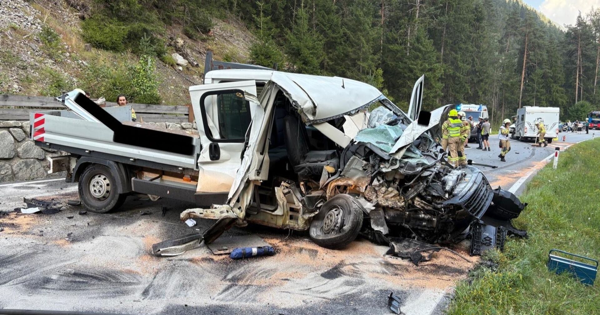 Schwer beschädigtes Firmenfahrzeug nach einem Frontalcrash im Ötztal. Die Fahrerkabine ist stark eingedrückt, Wrackteile liegen auf der Straße. Im Hintergrund arbeiten Feuerwehrleute und Einsatzkräfte an der Unfallstelle, ein Wohnmobil ist ebenfalls zu sehen.