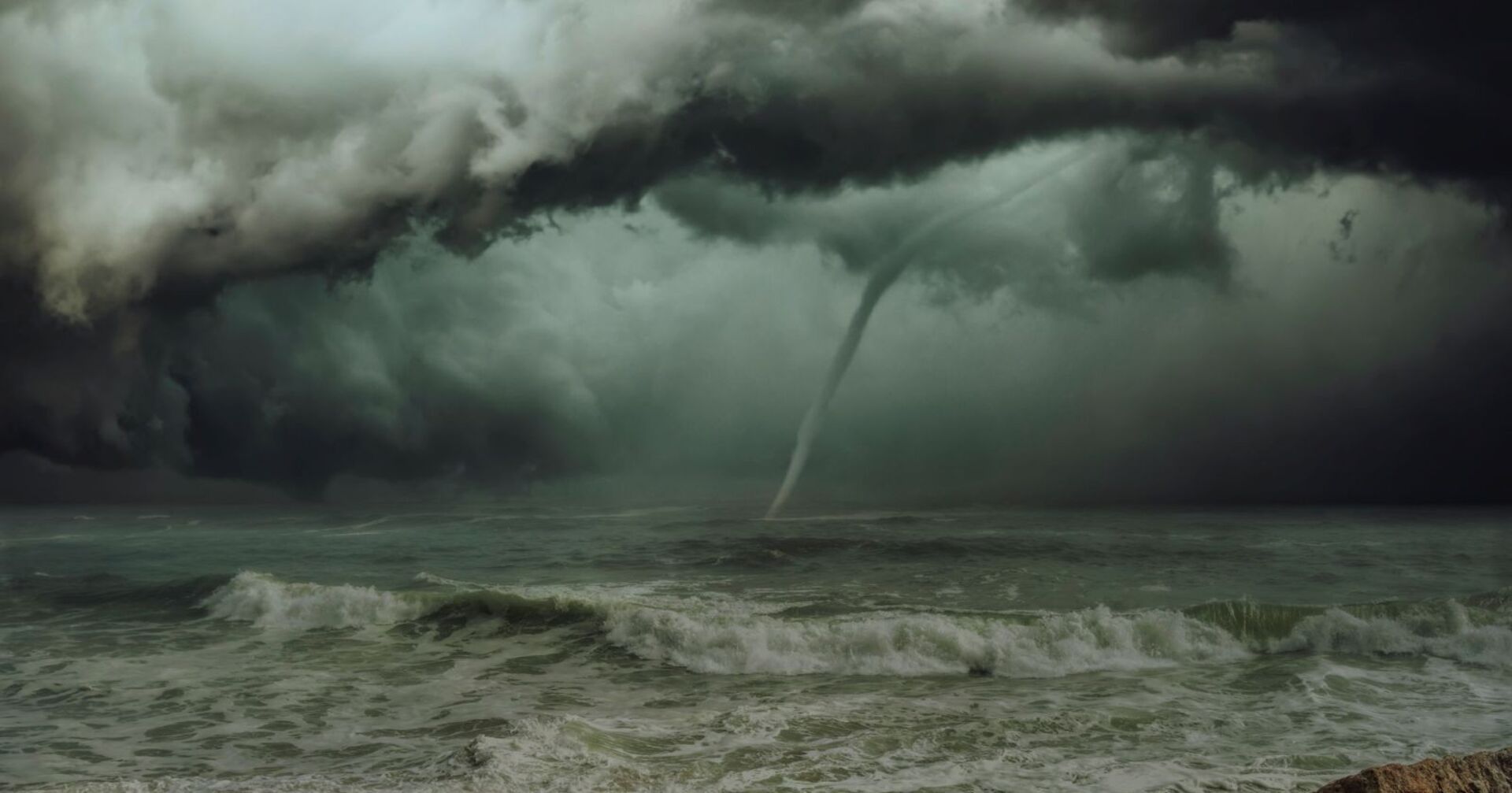 Dunkle Gewitterwolken über dem Meer mit einem gut sichtbaren Tornado, der sich aus der Wolkendecke bis zur Wasseroberfläche erstreckt. Im Vordergrund tobt die Brandung.