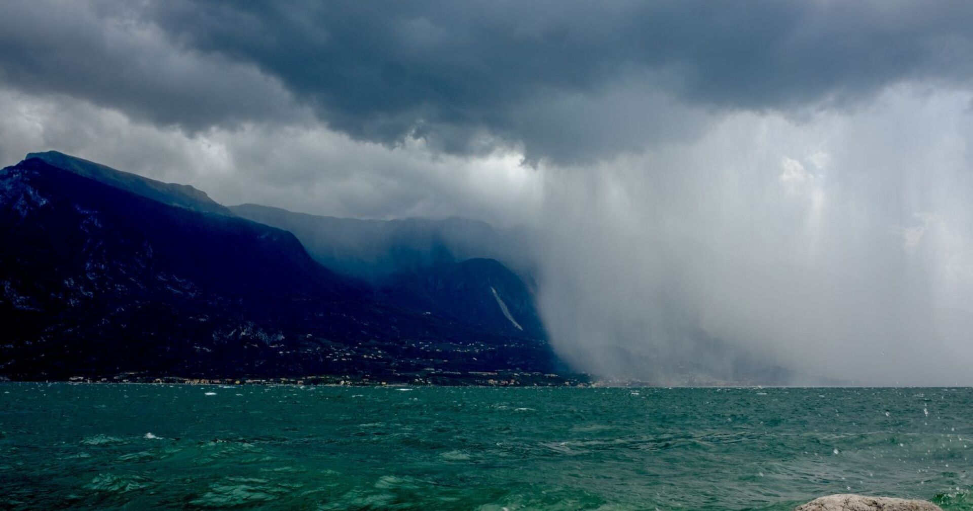 Dunkle Gewitterwolken und heftiger Starkregen über dem Gardasee. Der linke Bildteil zeigt dunkle Berge unter einer bedrohlichen Wolkendecke, rechts fällt dichter Regen über die Küste. Im Vordergrund ist aufgewühltes, türkisgrünes Wasser zu sehen.