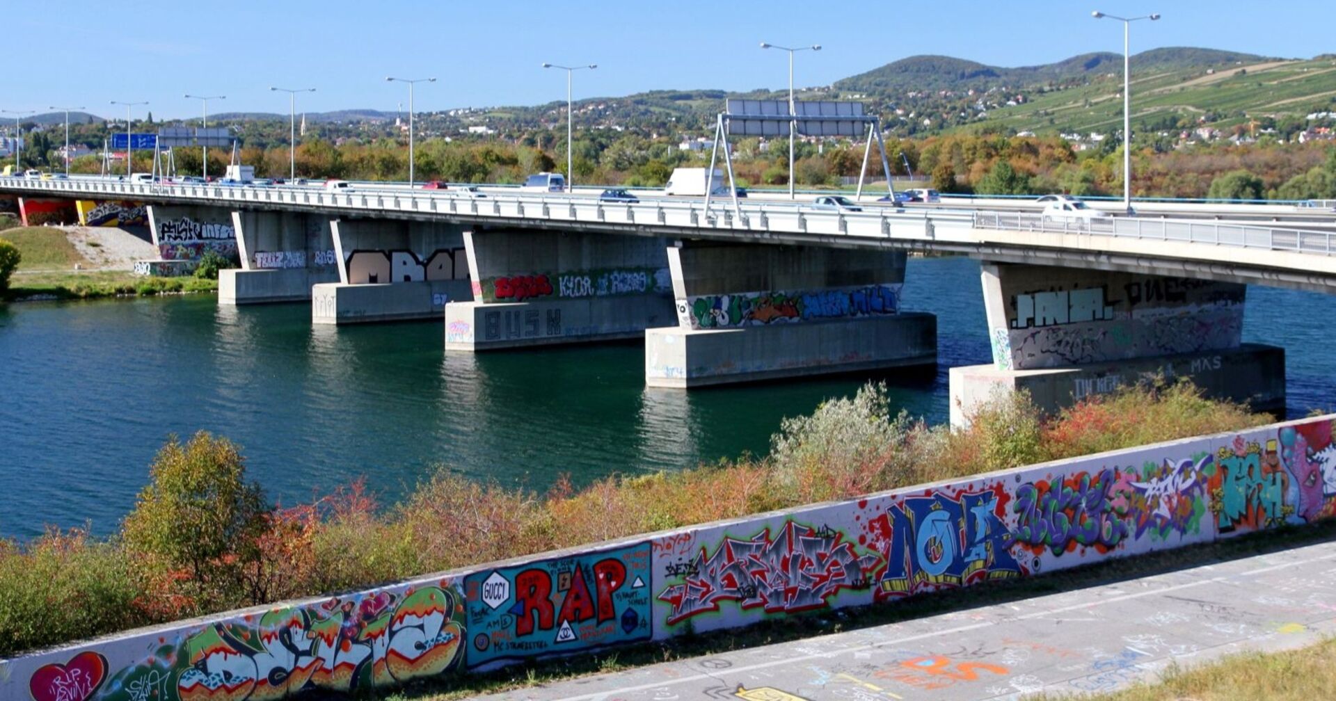 Blick auf eine Brücke über die Neue Donau in Wien, mit bunten Graffiti an den Brückenpfeilern und Mauern im Vordergrund. Im Wasser unterhalb der Brücke wurde eine Leiche gefunden.
