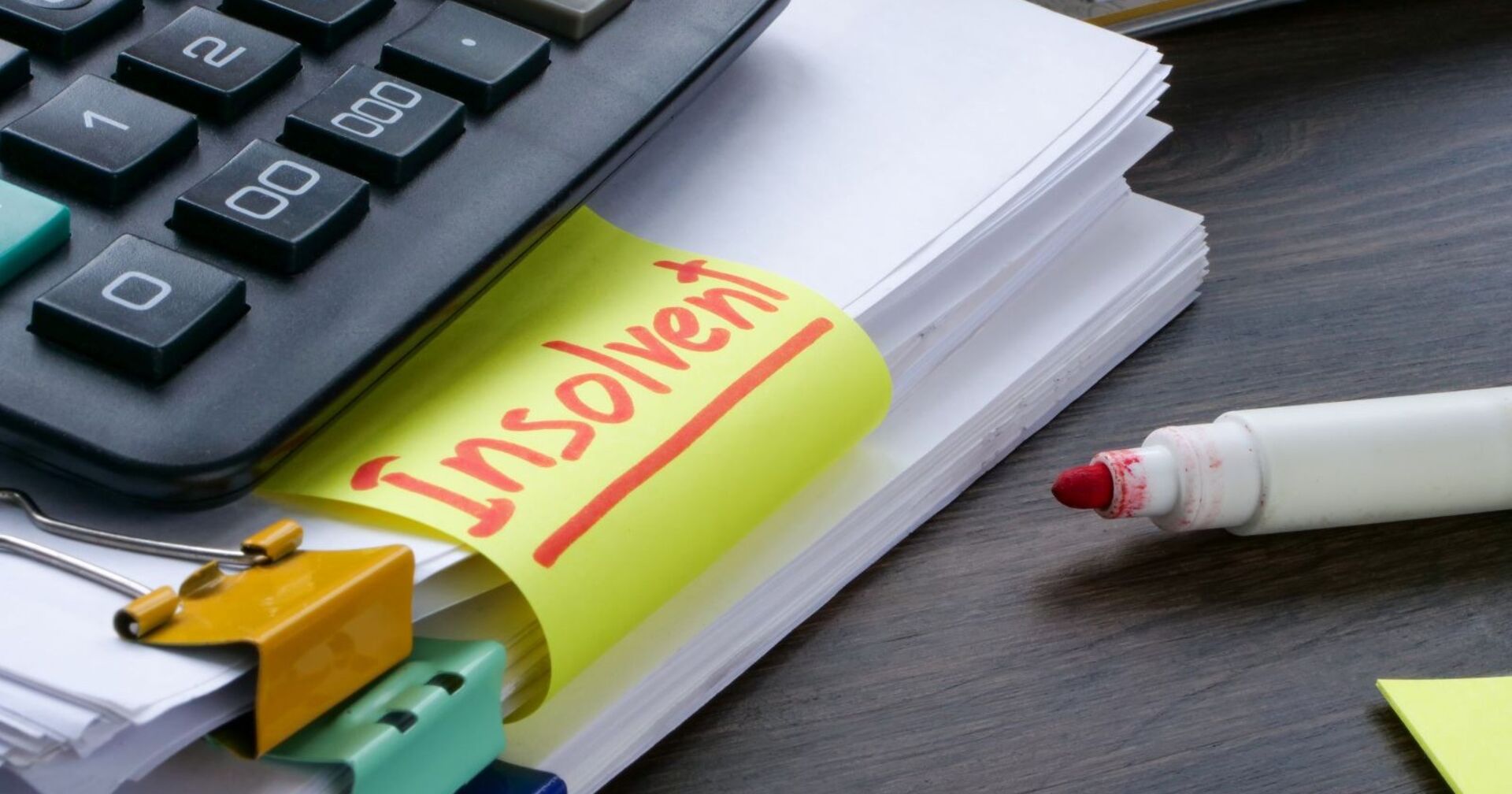 Bürotisch auf dem ein Stapel Papier mit einem Rotstift und ein Taschenrechner, auf einem Post-it steht das Wort insolvent geschrieben