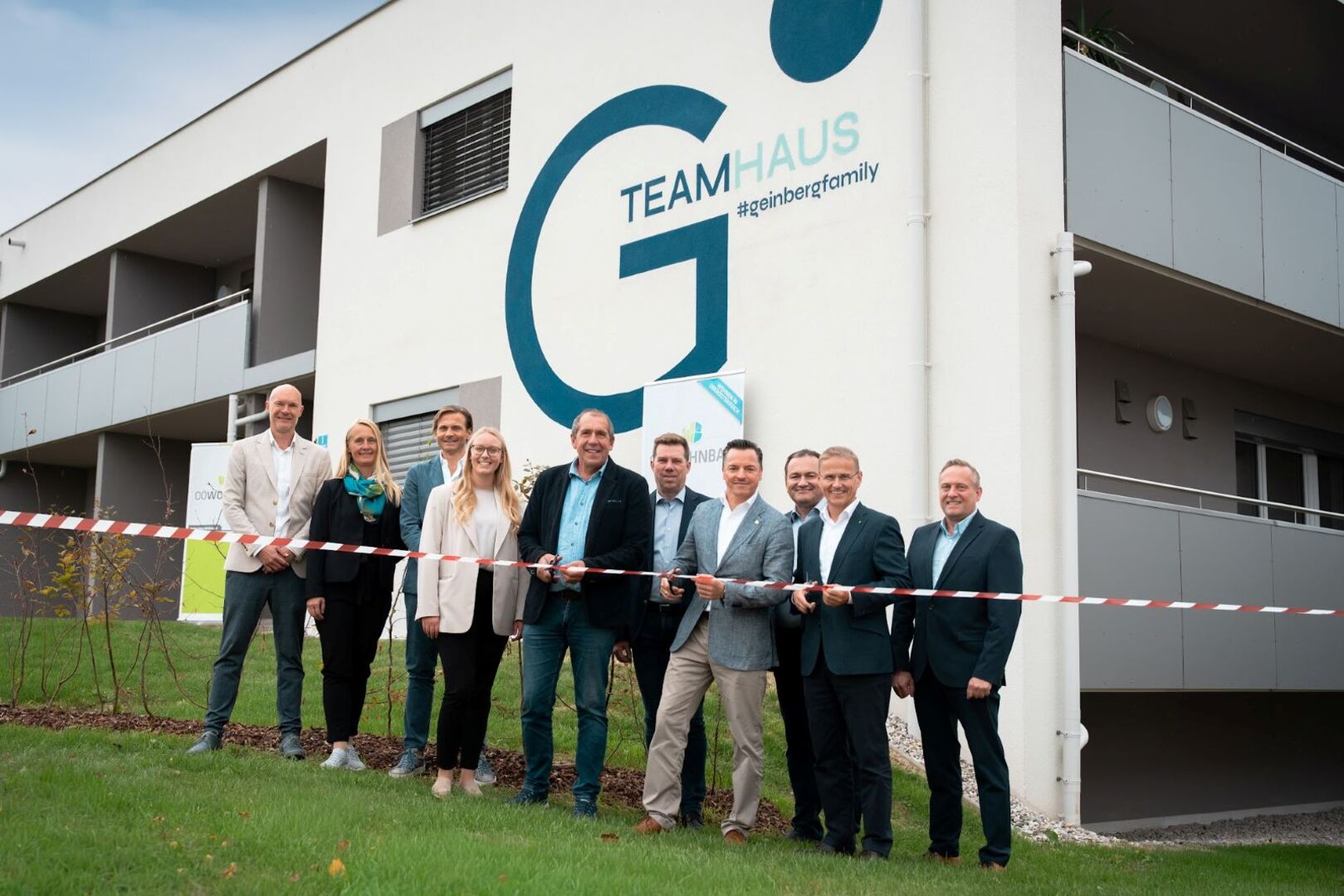 Zu sehen ist das neue Teamhaus für die Mitarbeiter des Spa Resort Geinberg. Im Vordergrund eine Gruppe von Menschen, die sich über die Eröffnung freuen. Einer der Anwesenden durschneidet ein rotes Band.