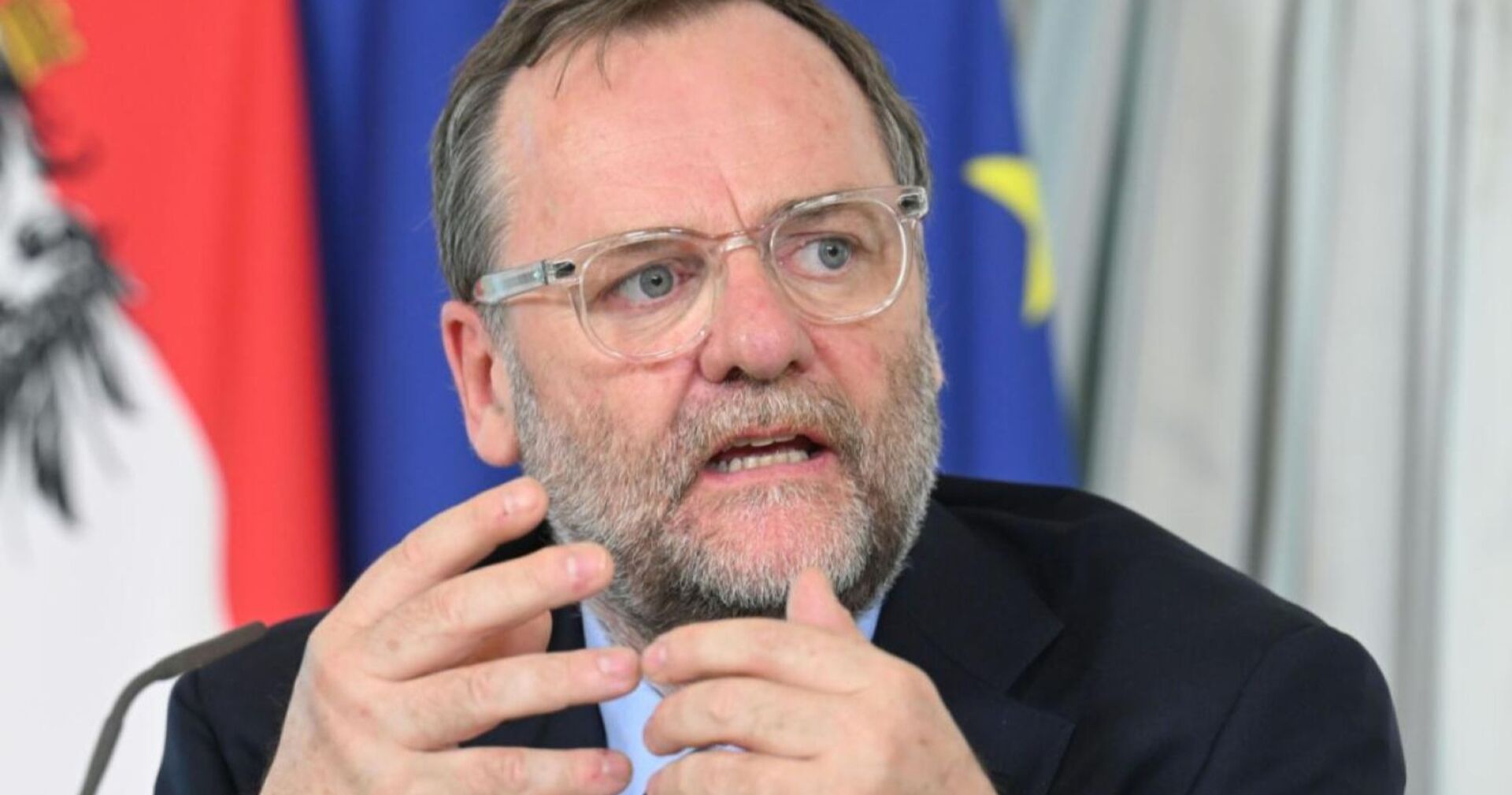 Sepp Schellhorn bei einer Pressekonferenz, sprechend mit Brille, vor der österreichischen und der europäischen Flagge.