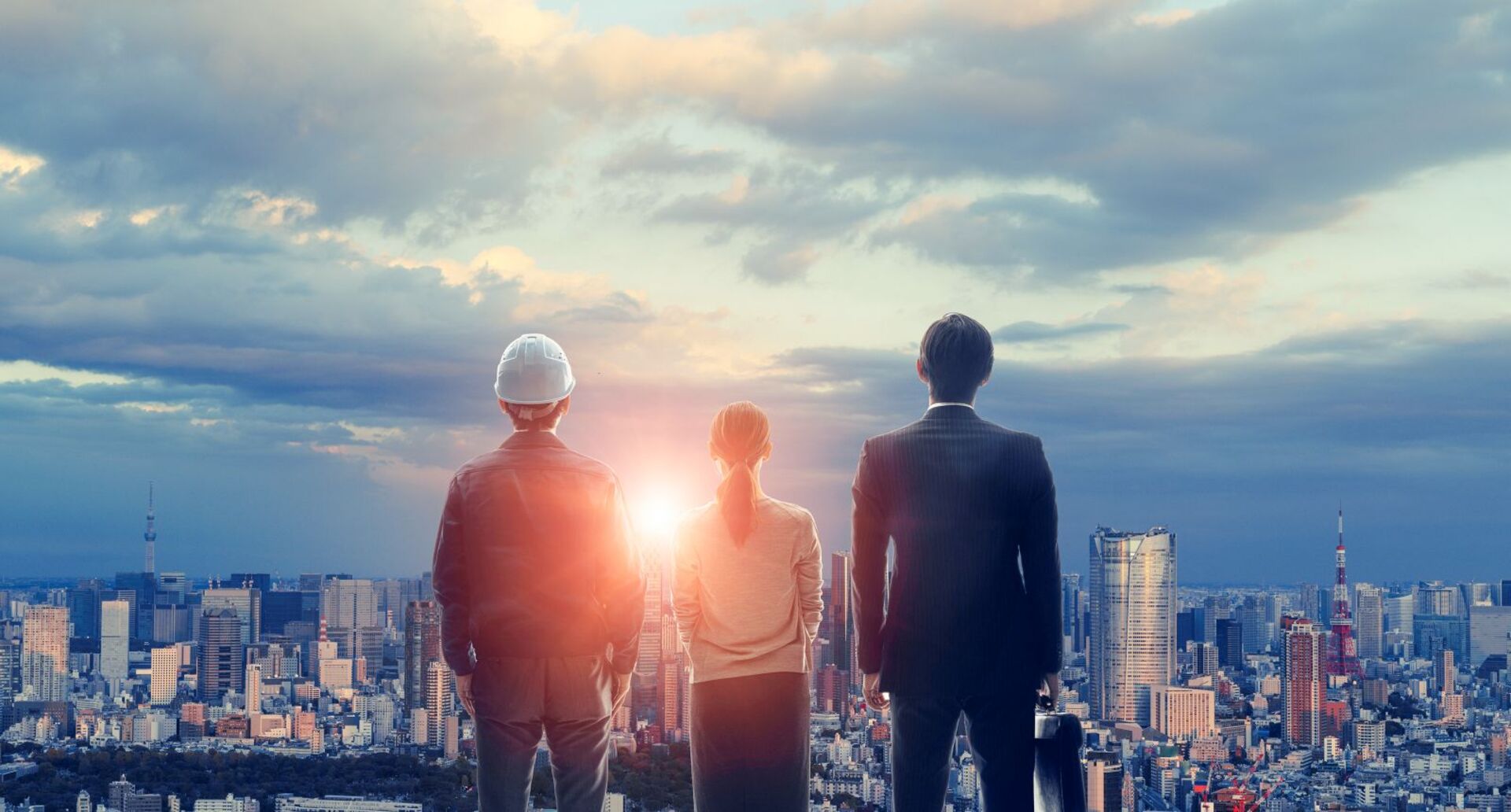 Ein Bauarbeiter, eine Frau und ein Geschäftsmann stehen sind von hinten zu sehen, vor ihnen liegt eine große Stadt, in der gerade die Sonne aufgeht.