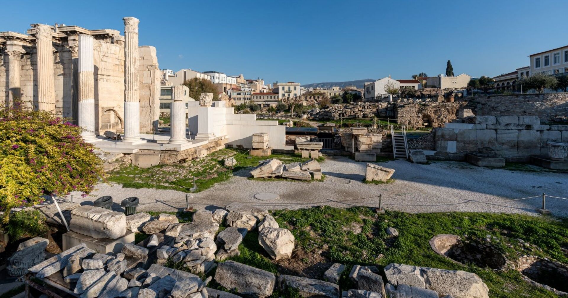 Ruinen mit Säulen und Steinresten in Athen, antike Überreste vor moderner Stadtkulisse bei klarem Himmel.