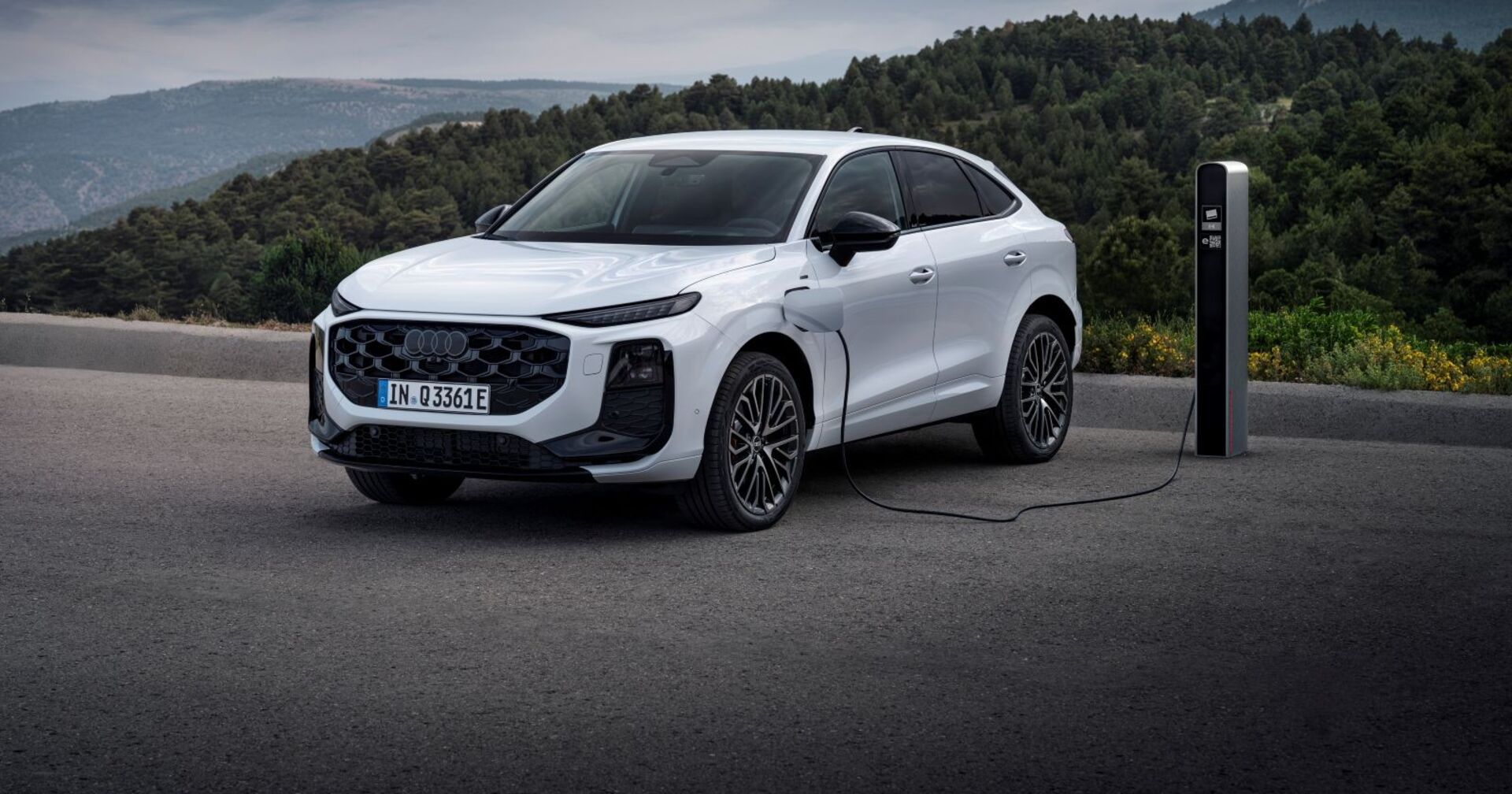 Ein weißer Audi Sportback an einer Ladestation. Das Auto parkt auf einem asphaltierten Parkplatz, im Hintergrund sind bewaldete Hügel zu sehen.
