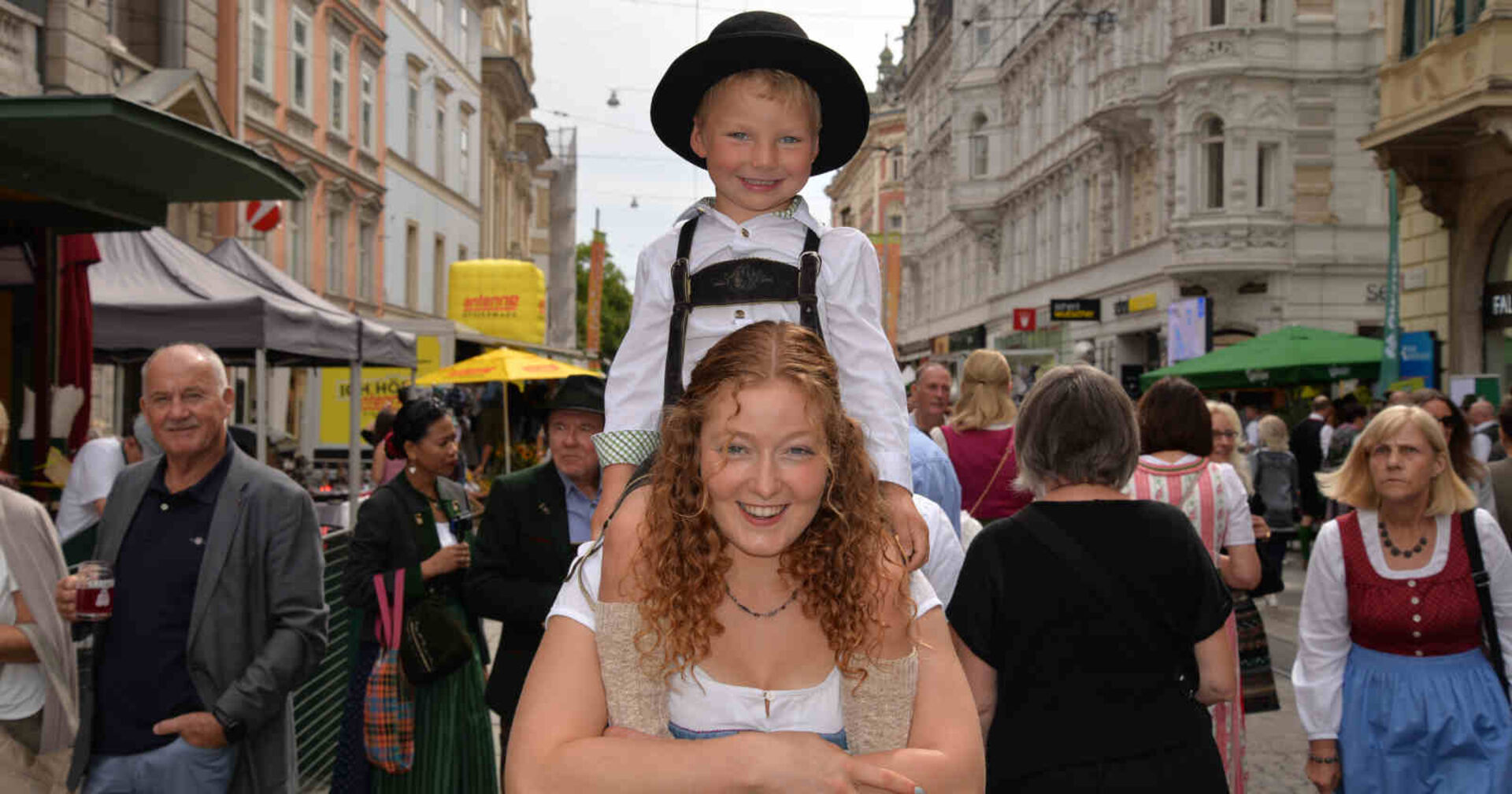 Ein kleiner Bub sitzt auf den Schultern eines rothaarigen Mädchens