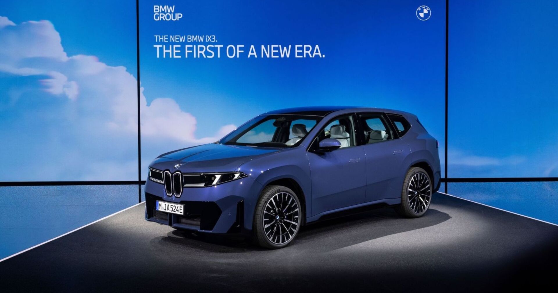 Ein blauer BMW IX3 steht in einem Präsentationsraum vom BMW. Im Hintergrund ist eine blaue Wand mit einigen Wolken zu sehen.