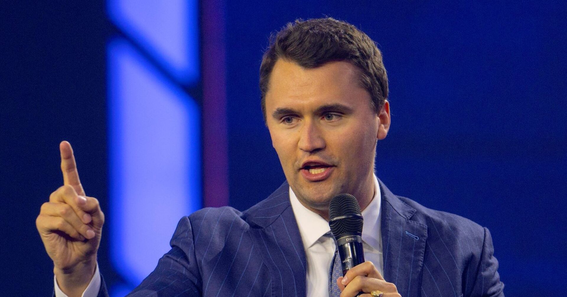 Charlie Kirk im Anzug mit Mikrofon, er hebt den Finger und spricht während eines öffentlichen Auftritts.