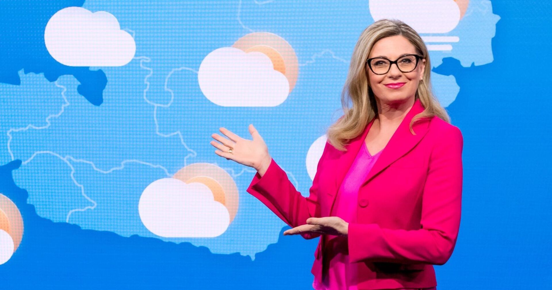 Christa Kummer in pinkem Blazer vor einer Wetterkarte mit Wolken-Symbolen – sie zeigt mit der Hand auf die Grafik und lächelt in die Kamera.