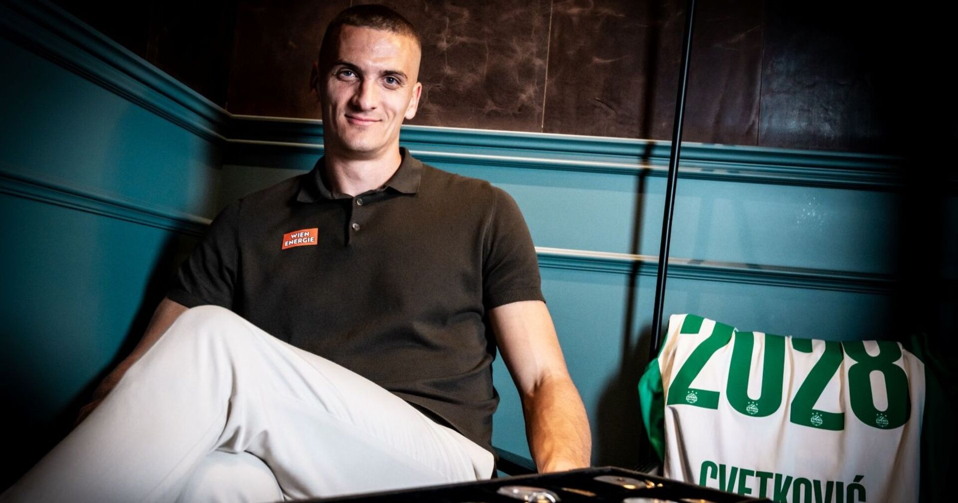 Nenad Cvetkovic in einem Poloshirt mit einem Sticker des Sponsors Wien Energie, im Hintergrund ein Rapid-Trikot, welches das jahr seines weiteren vertrages zeigt