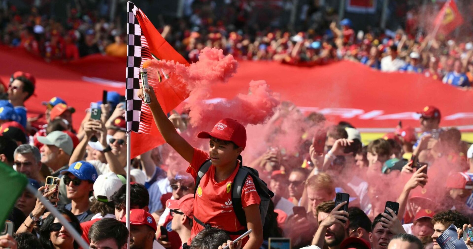 Ein Junge im Ferrari-Outfit hält eine rot-weiß karierte Fahne und eine rote Rauchfackel inmitten einer jubelnden Menschenmenge beim Formel-1-Rennen in Monza.