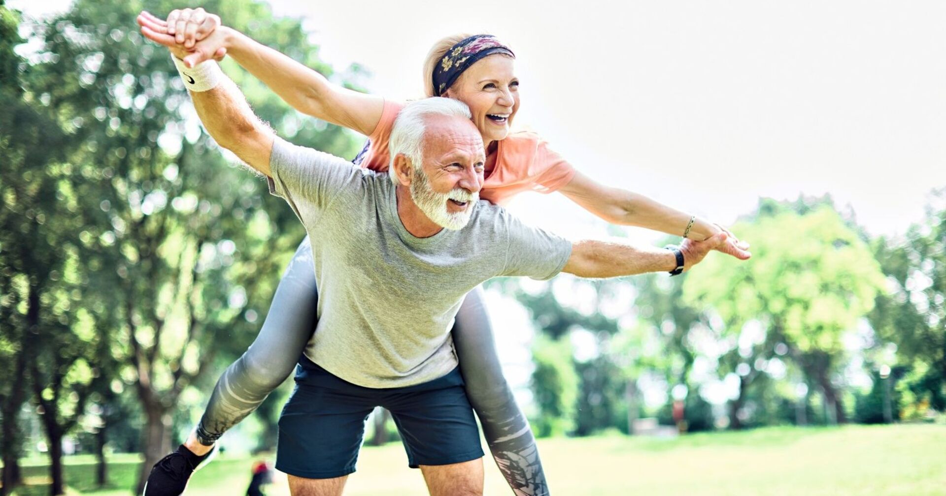 Ein Mann und eine Frau machen Fitnessübungen im Grünen. Er hat weiße Haare und einen weißen Bart und trägt ein graues T-Shirt sowie eine kurze, blaue Hose. Die Dame umklammert ihn von hinten und lacht. Sie trägt eine rosa T-Shirt und eine silberne, lange Trainingshose. Ihr Haar ist mit einem blauen Band zusammengehalten. Im Hintergrund sieht man einige Laubäume.