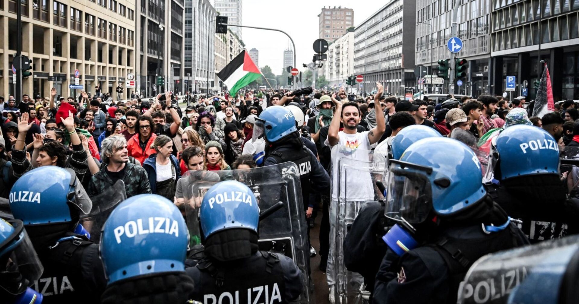 Menschenmenge mit palästinensischen Fahnen protestiert auf einer Straße in Italien, während eine Reihe von Polizisten in blauer Schutzausrüstung und Helmen den Weg versperrt.