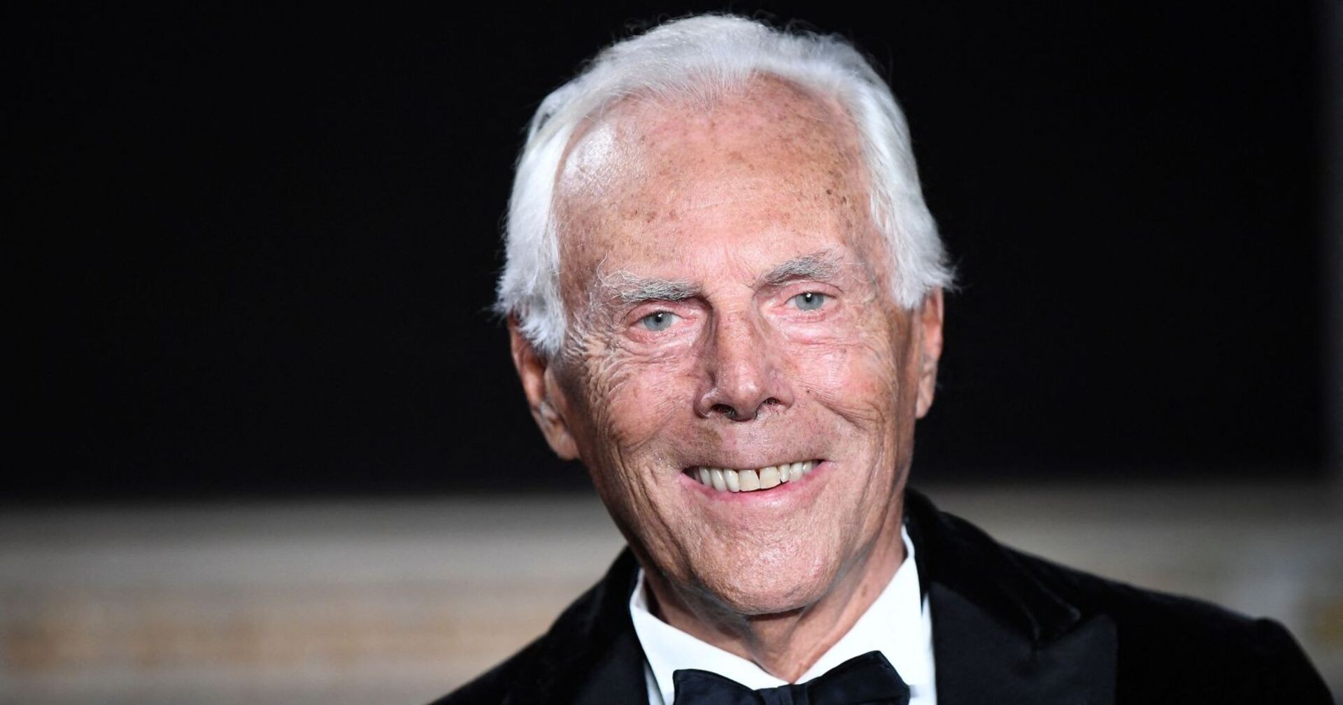Porträt von Giorgio Armani mit weißem Haar, der im Smoking und mit Fliege in die Kamera lächelt.