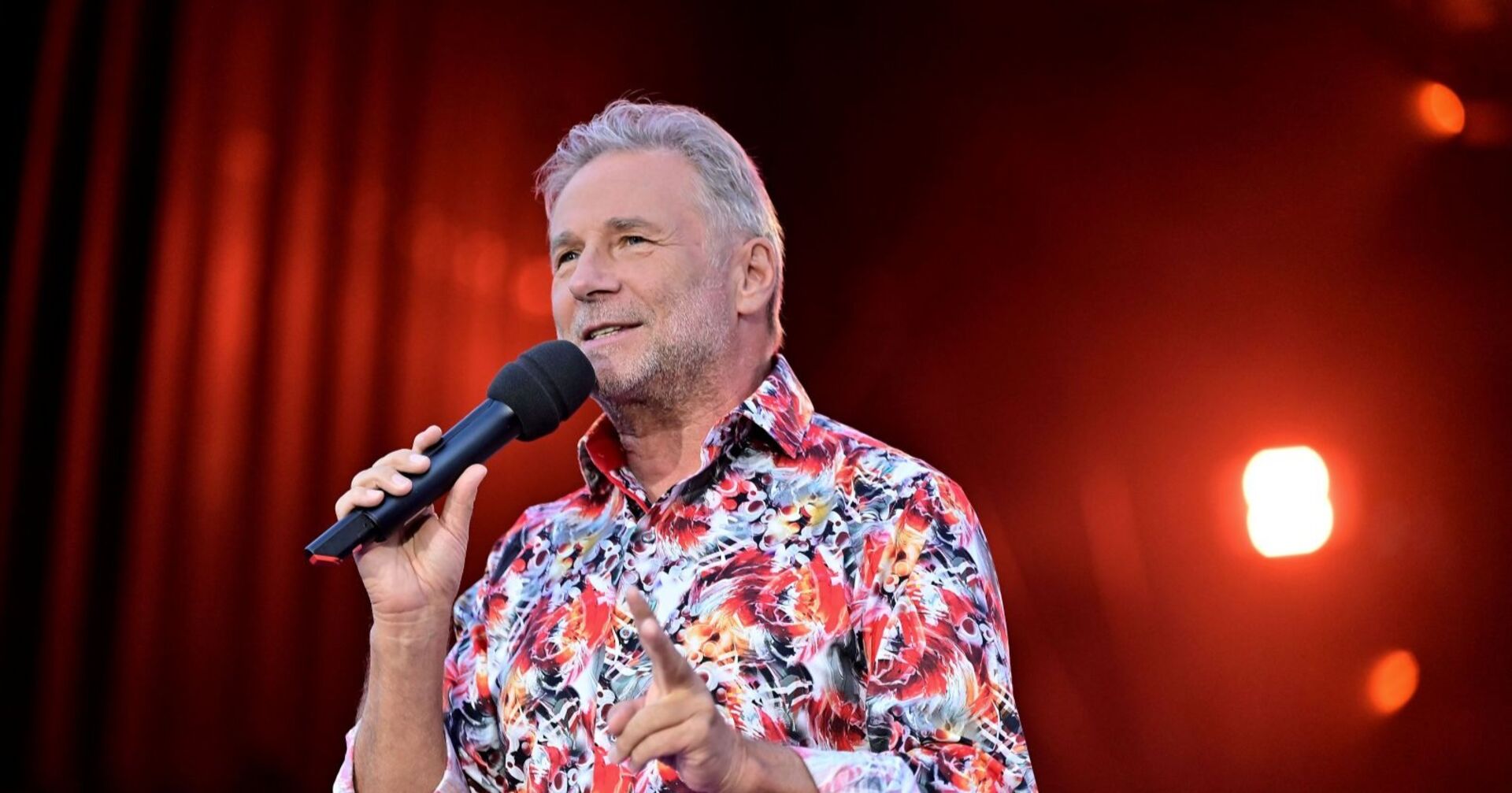 Friedl Würcher, Frontmann der Nockis, mit Mikrofon auf der Bühne in einem bunt gemusterten Hemd vor rotem Hintergrund.