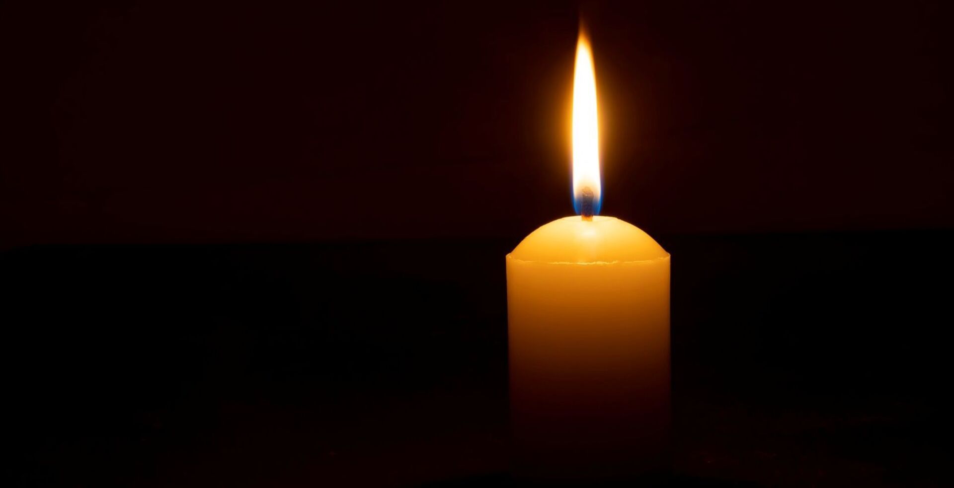 Einzelne brennende Kerzenflamme oder Licht, das auf einer großen weißen Kerze auf schwarzem oder dunklem Hintergrund auf dem Tisch in der Kirche für Weihnachten, Beerdigung oder Gedenkfeier mit Kopierraum leuchtet.