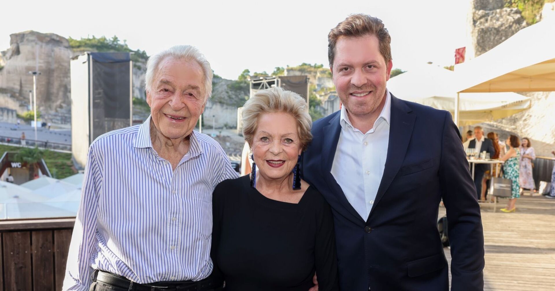 Harald Serafin steht lächelnd neben seiner Frau Ingeborg und Sohn Daniel. Im Hintergrund ist die Felsenbühne der Oper im Steinbruch St. Margarethen zu sehen.