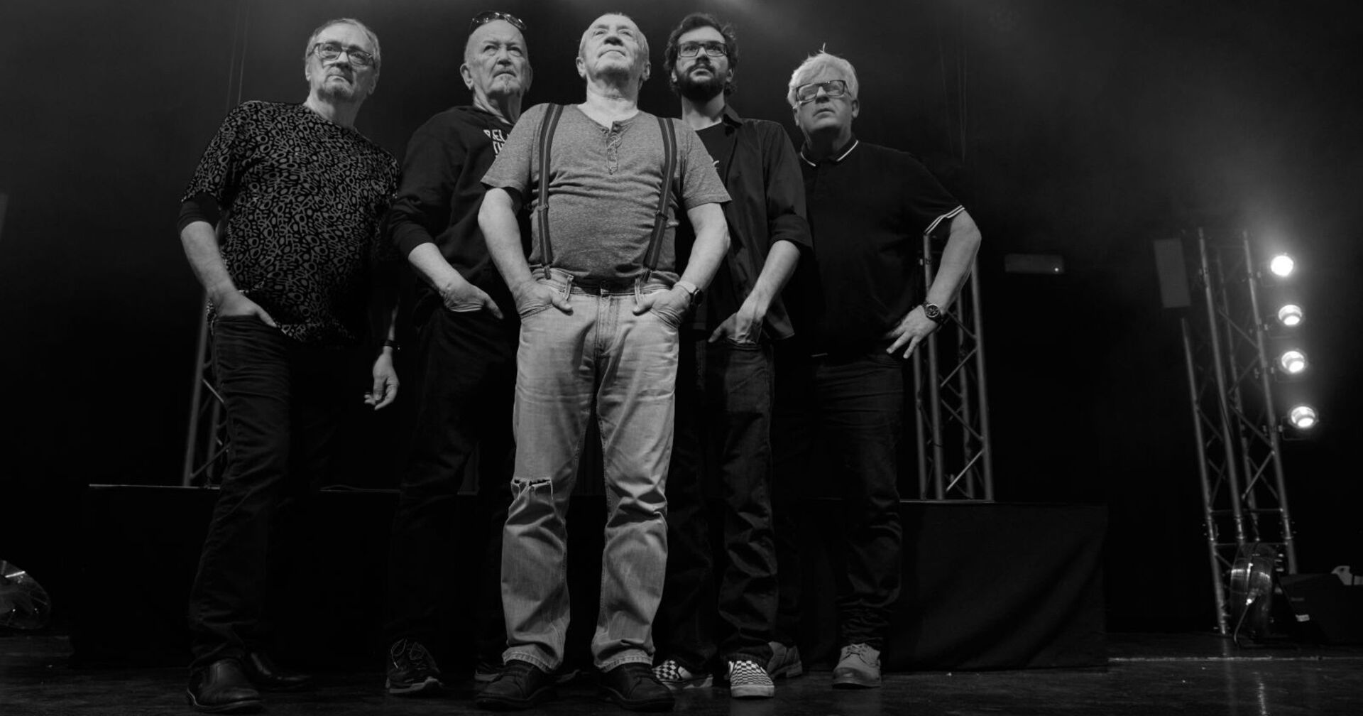Schwarz-Weiß-Foto einer fünfköpfigen Band auf der Bühne, die nebeneinander in die Kamera blickt. Die Musiker tragen legere Kleidung und stehen vor einer Bühnenkulisse mit Scheinwerfern.
