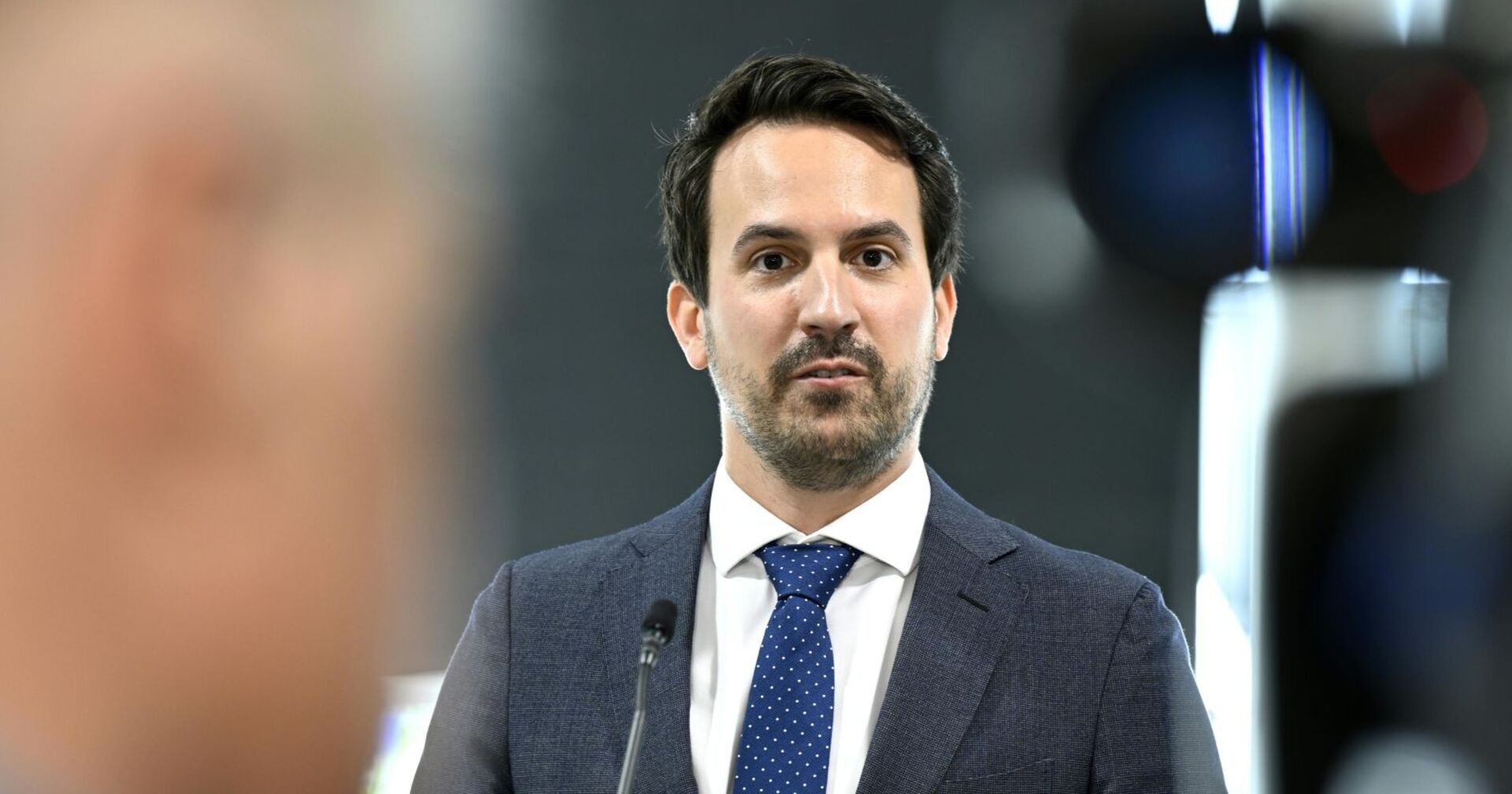 Christoph Wiederkehr spricht bei einer Pressekonferenz; der Bildungsminister trägt Anzug und Krawatte, im Vordergrund unscharf eine Person und Kameraequipment.