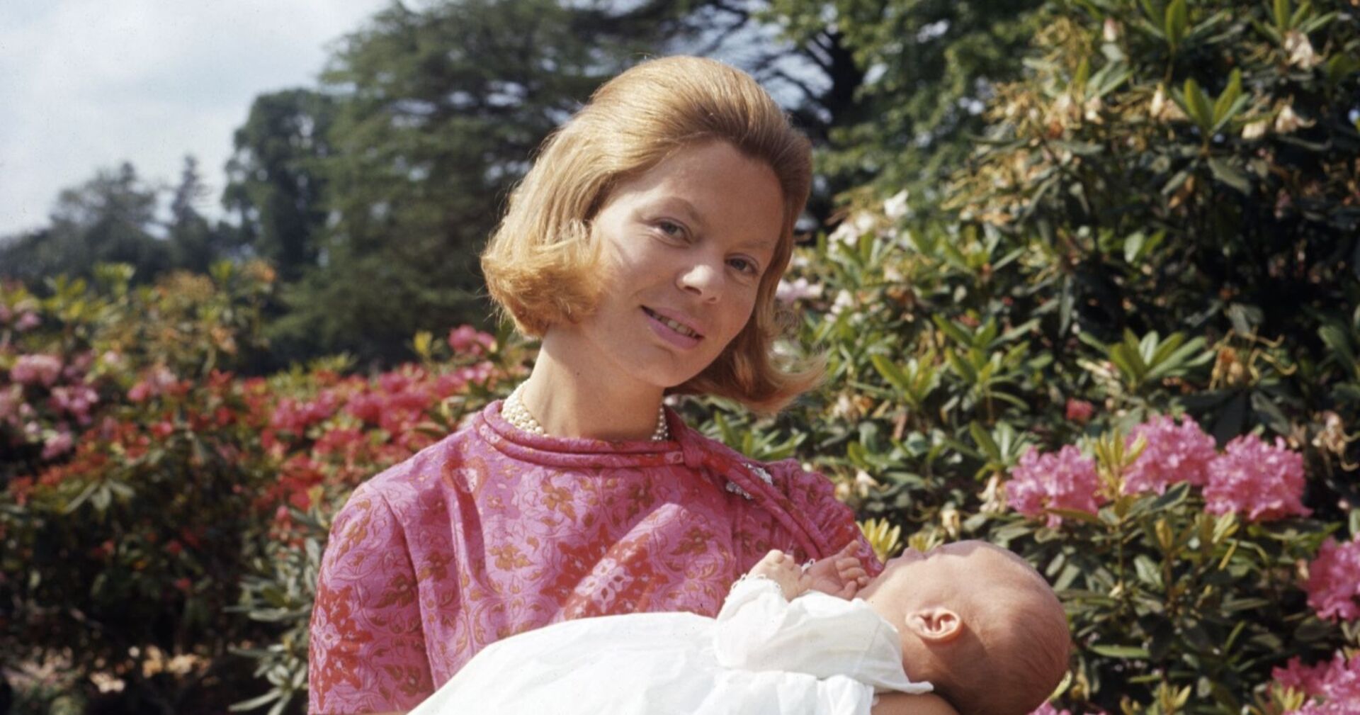 Junge Frau in rosa Kleid mit Perlenkette hält ein Neugeborenes im Arm, im Hintergrund blühende Sträucher und Bäume.