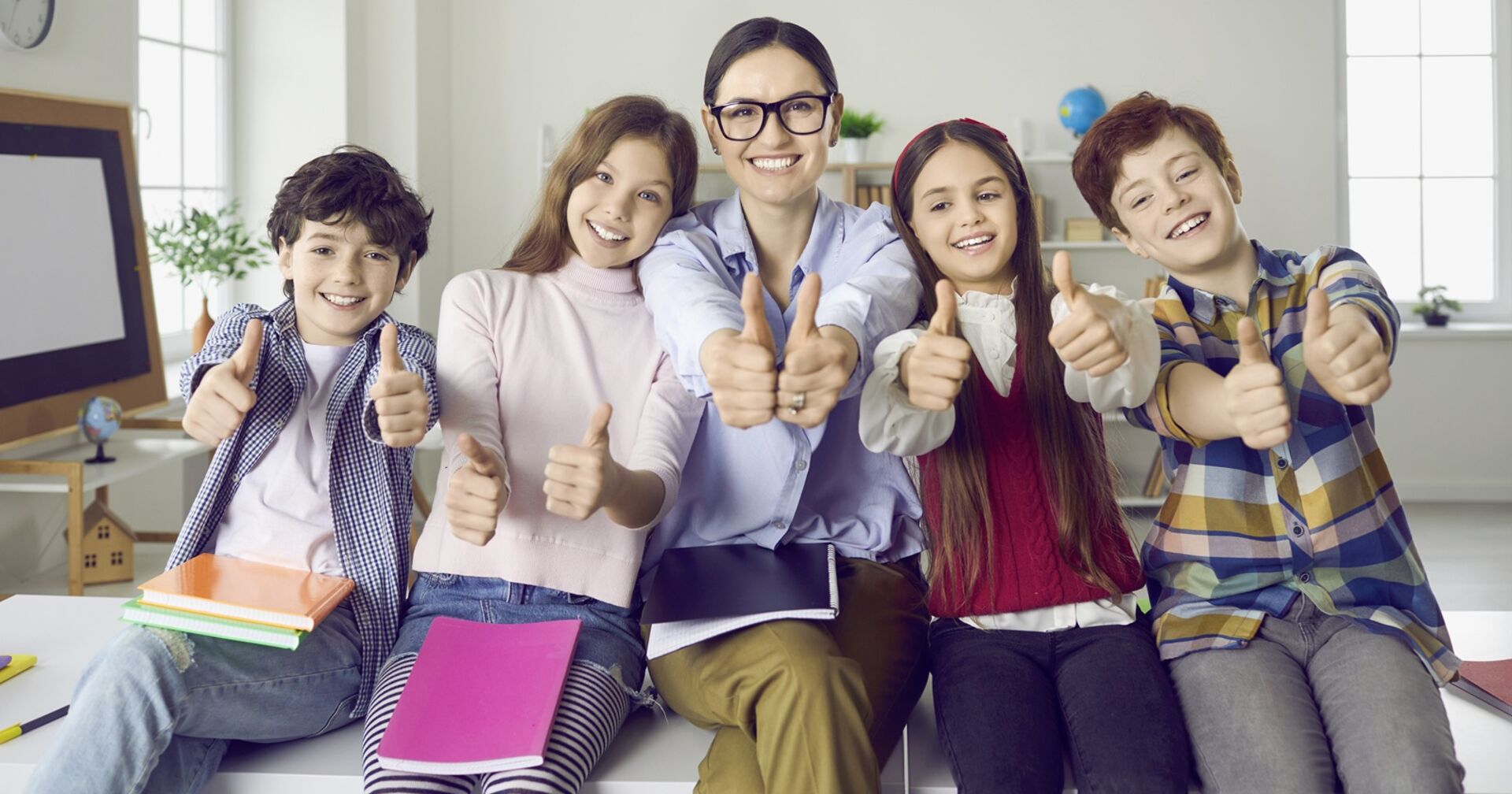 Eine junge Lehrerin und vier Schülerinnen sitzen auf einem Tisch und zeigen den nach oben gestreckten Daumen