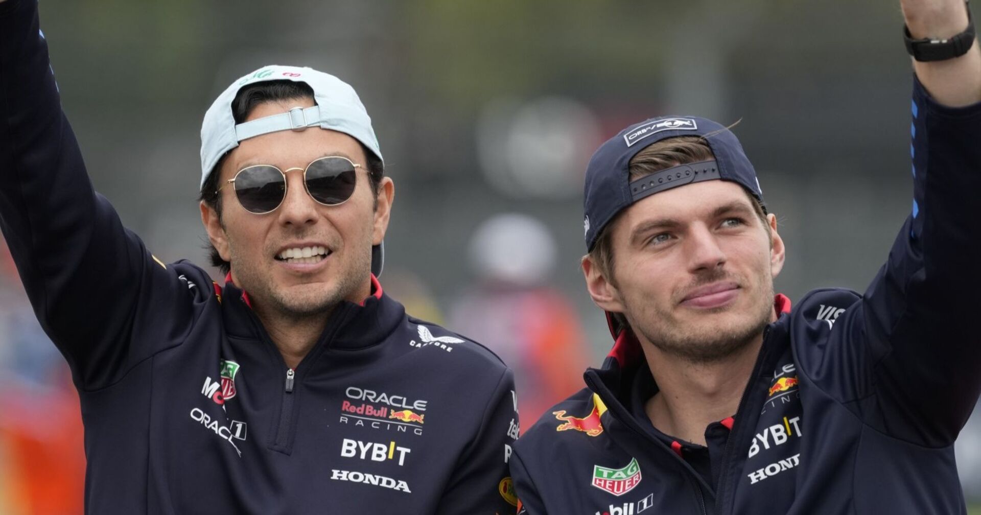 Sergio Pérez (links) und Max Verstappen (rechts) in Red-Bull-Teamkleidung, beide tragen Kappen und winken ins Publikum.