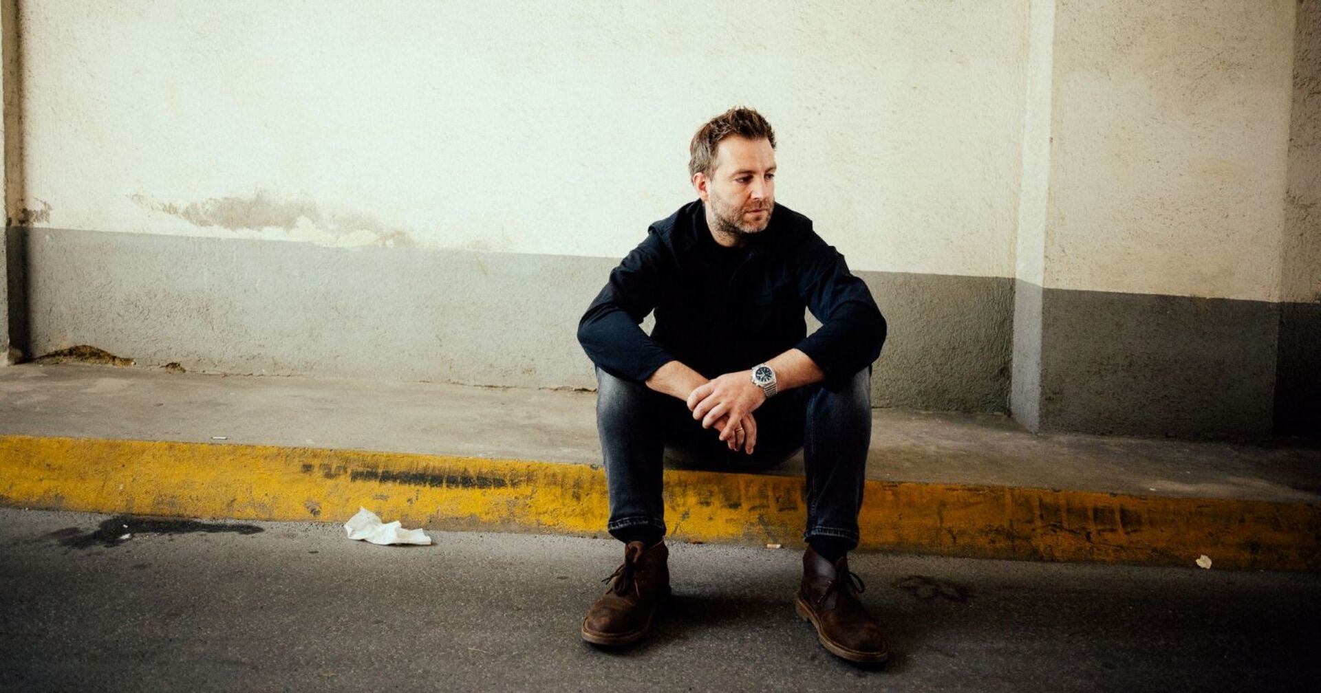 Josh. sitzt in einer Garageneinfahrt auf einem Randstein und sieht nachdenklich in die Ferne mit geneigtem Kopf